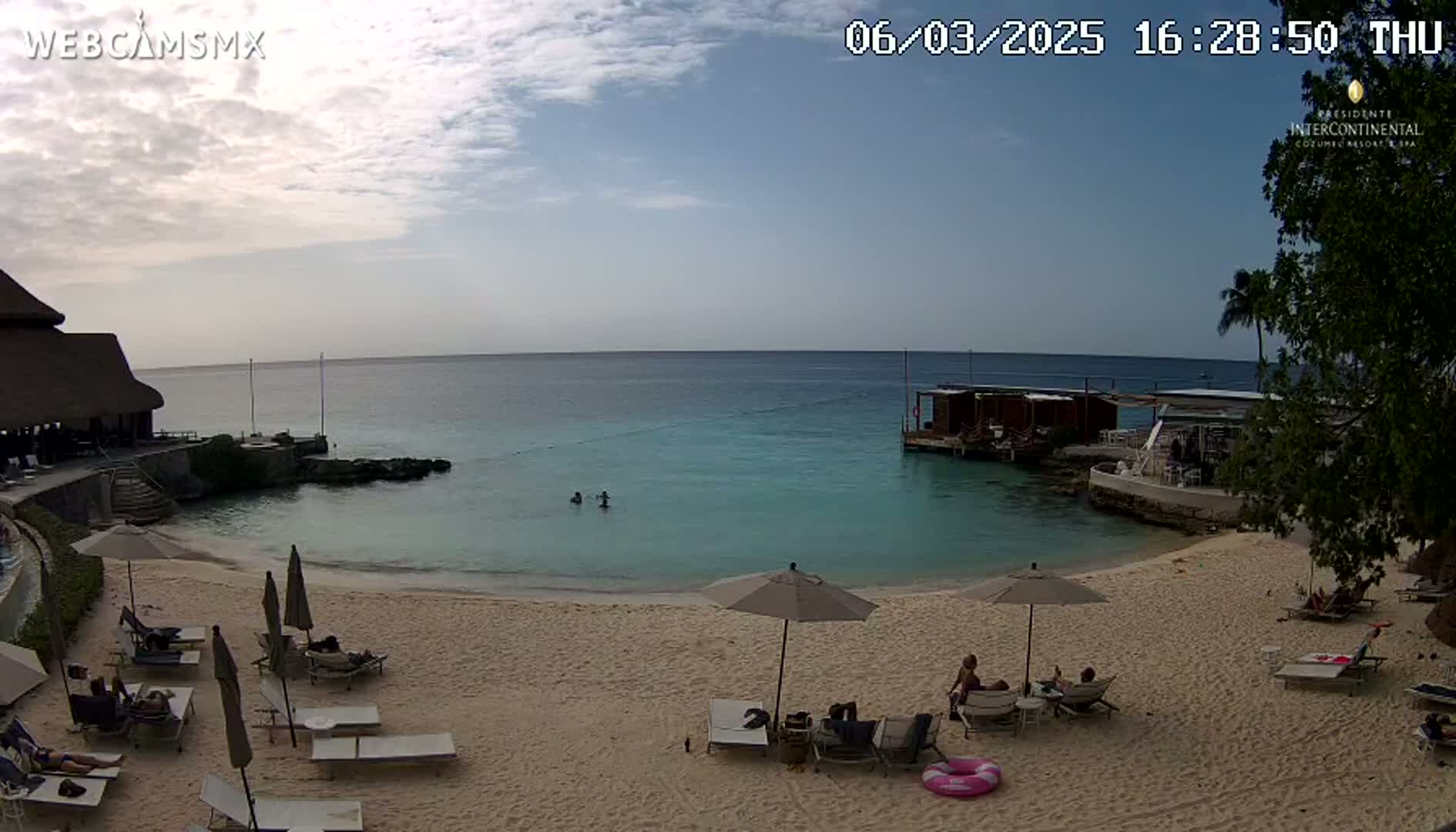 A sandy beach with lounge chairs and umbrellas under a partly cloudy sky, overlooking a calm, turquoise ocean with a few people swimming.