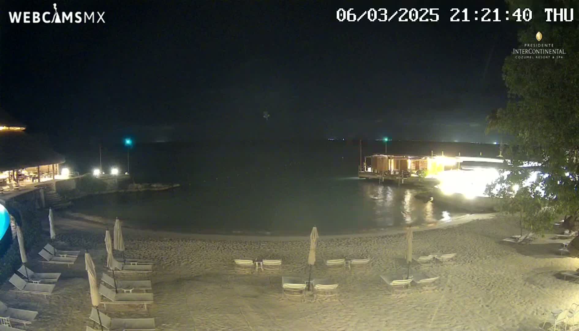 A nighttime beach scene shows a sandy shoreline with lounge chairs and umbrellas under a clear, dark sky, next to a calm body of water with lit structures along the edge.