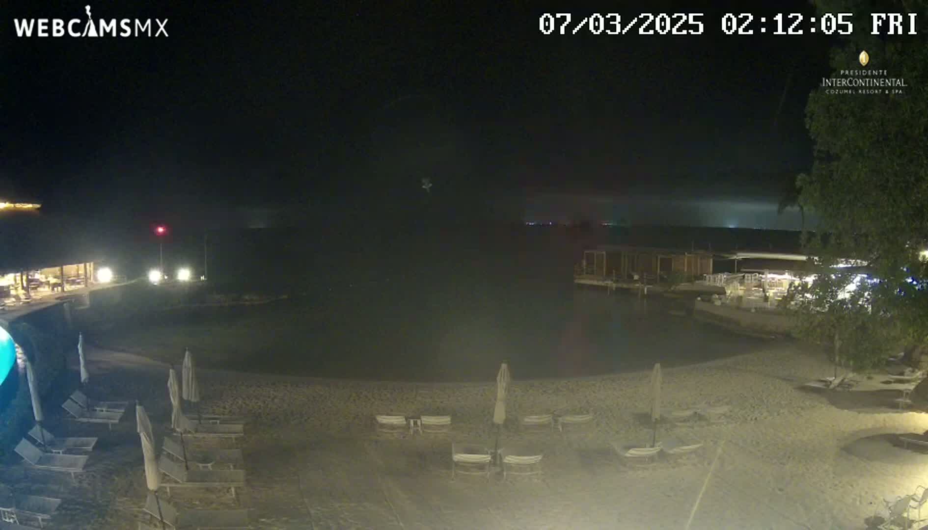 A nighttime beach scene shows a calm body of water, a sandy shoreline with empty lounge chairs and umbrellas, and some buildings with lights in the background.