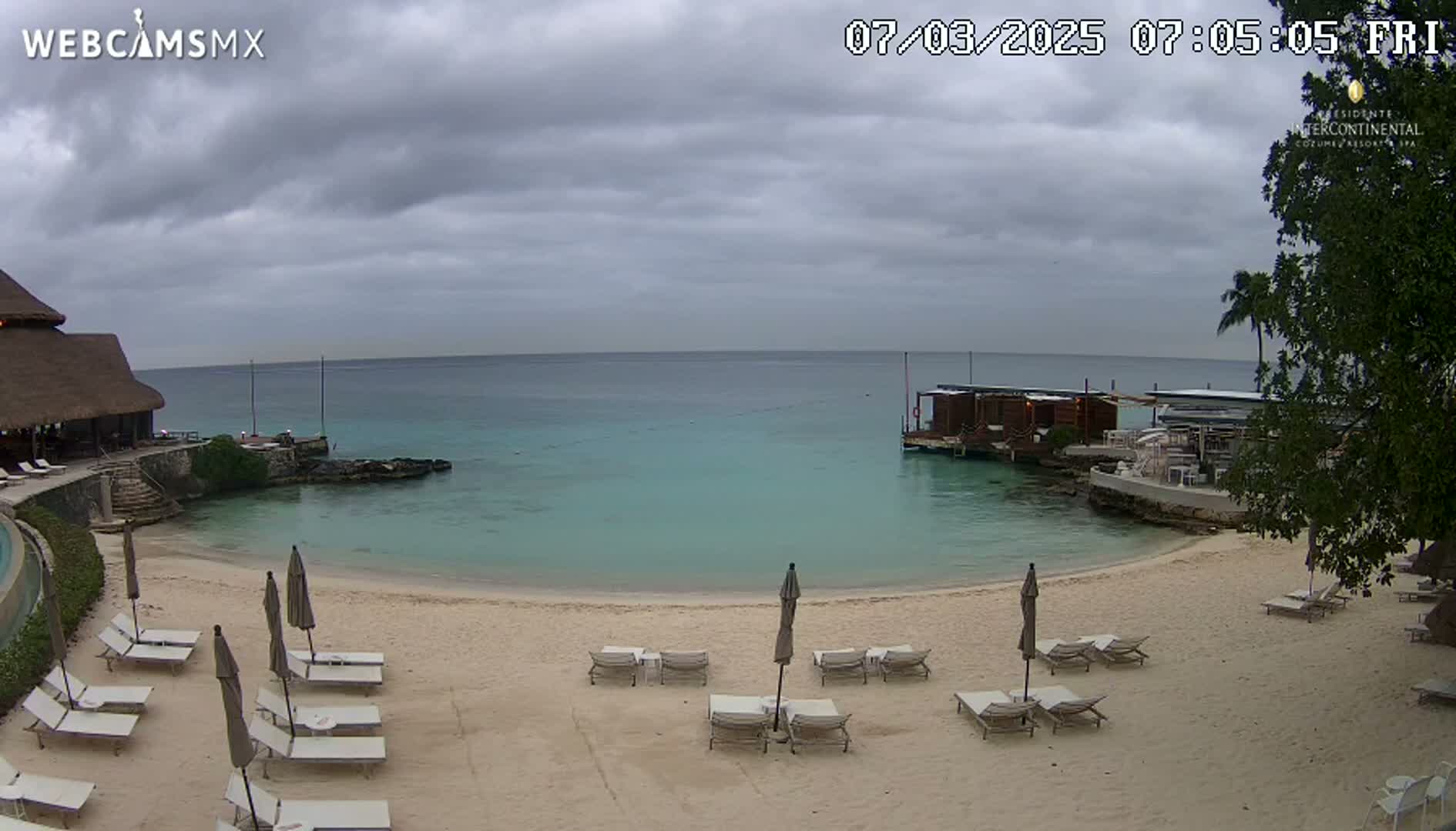 A sandy beach with lounge chairs and umbrellas sits in front of calm, turquoise water under a cloudy sky.