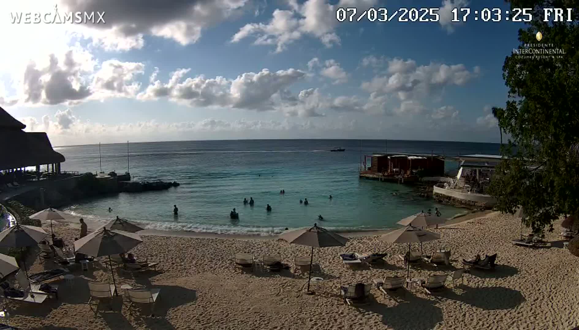 A partly cloudy day at a beach resort shows people swimming in calm, shallow water near a sandy beach with lounge chairs and umbrellas.
