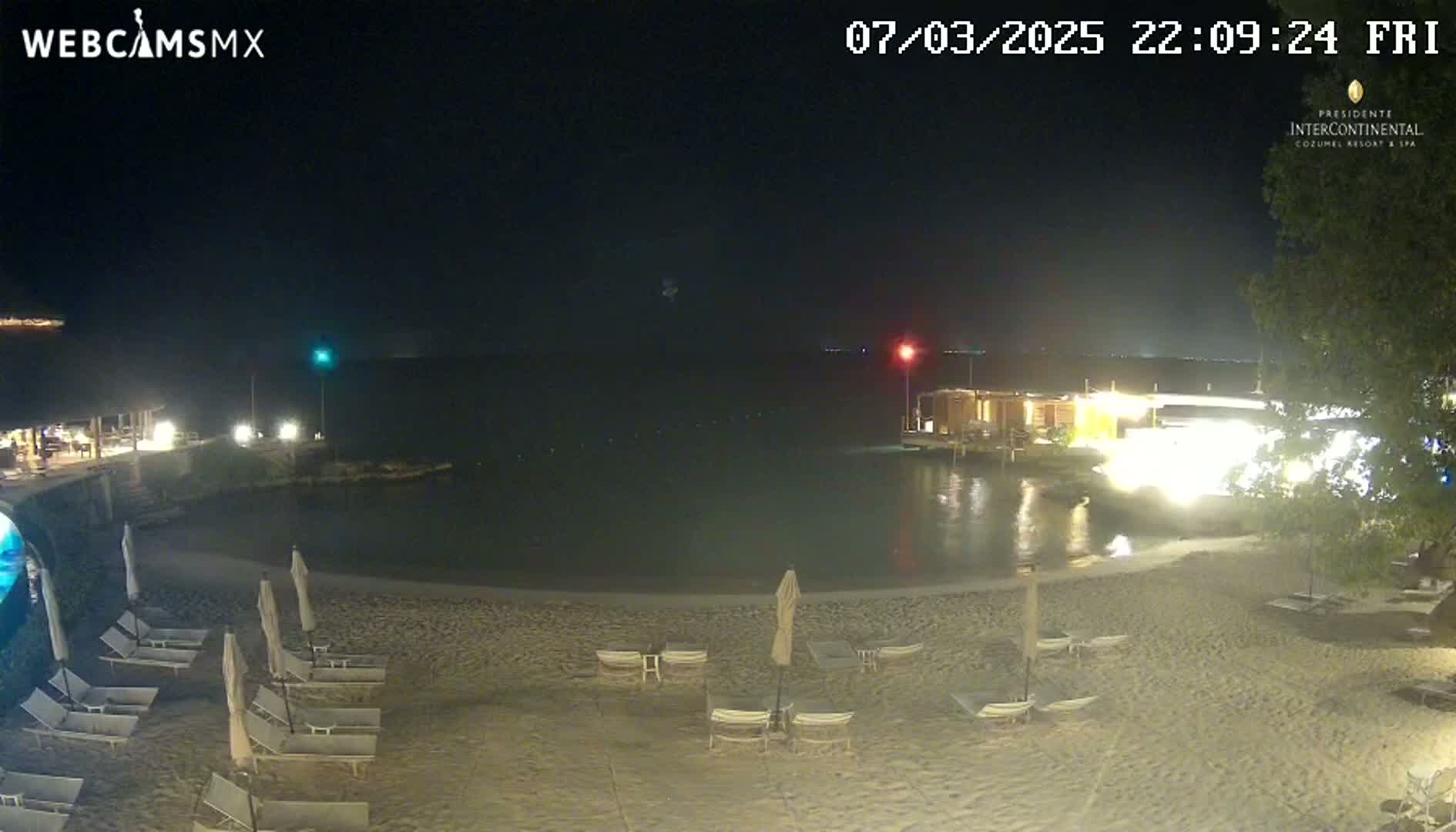 A sandy beach at night with lounge chairs and umbrellas is situated next to a calm body of water, with lights illuminating buildings in the background under a clear, dark sky.