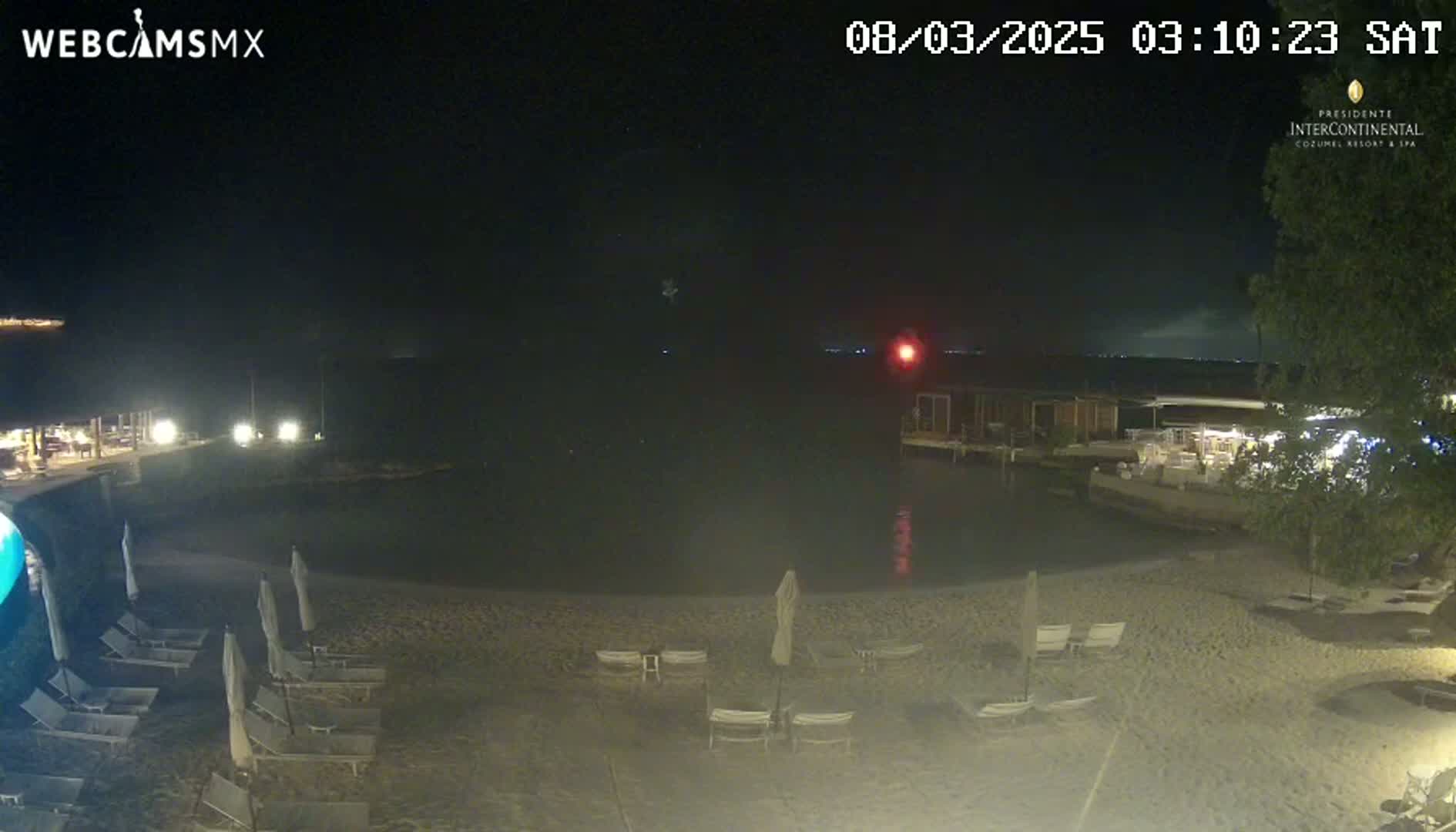 A nighttime view of a sandy beach with lounge chairs and umbrellas surrounding a calm body of water, next to a resort under a clear, dark sky.