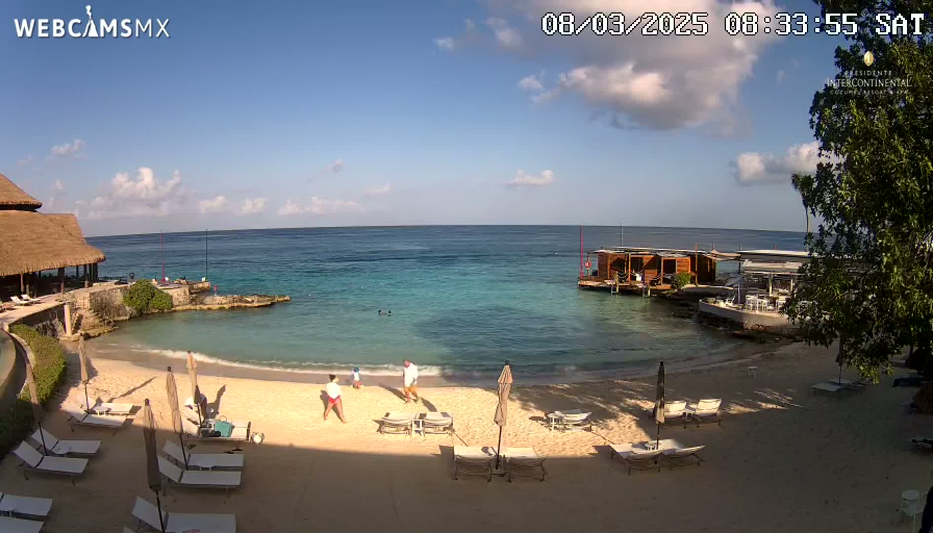 A sandy beach with lounge chairs and a few people is visible, with calm, clear turquoise water extending to a horizon under a partly cloudy sky.