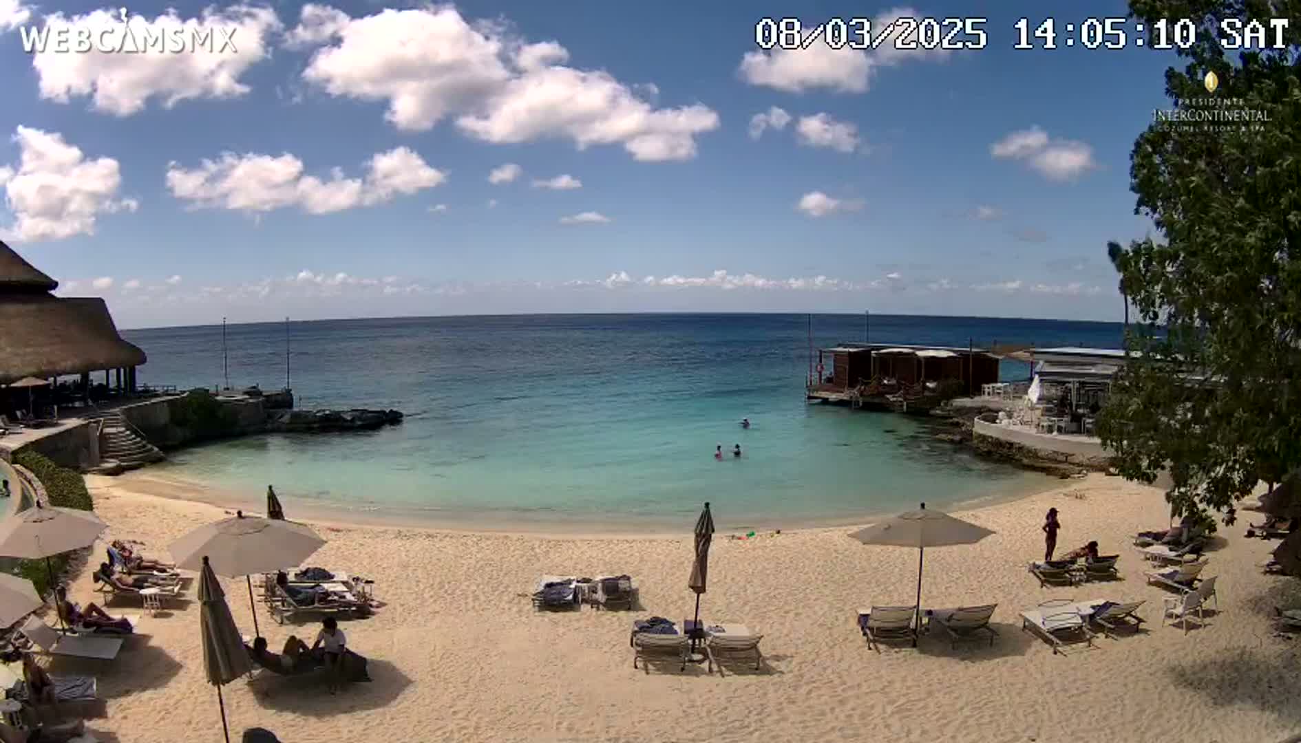 A sandy beach with turquoise water, beach umbrellas, lounge chairs, and a few people relaxing under a mostly sunny sky.