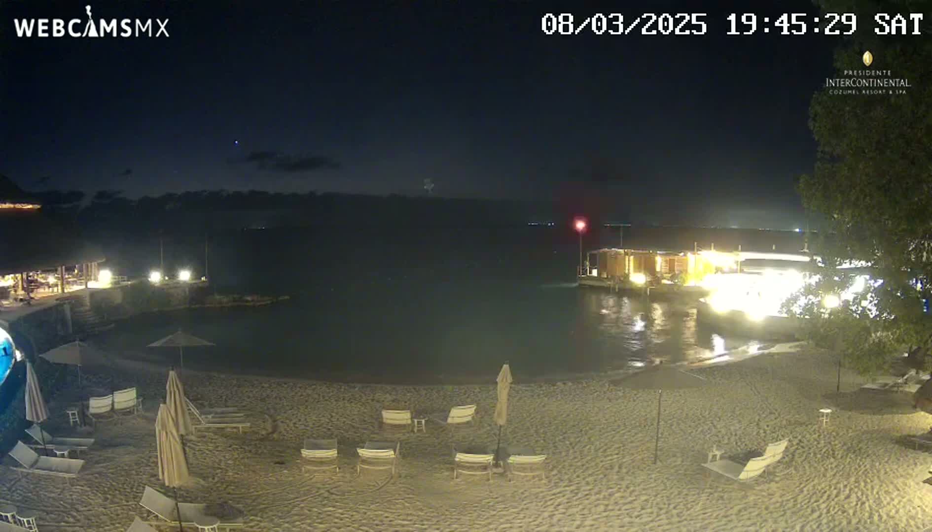 A nighttime beach scene shows a sandy shore with lounge chairs and umbrellas under a dark sky, with calm water and lit buildings visible in the background.