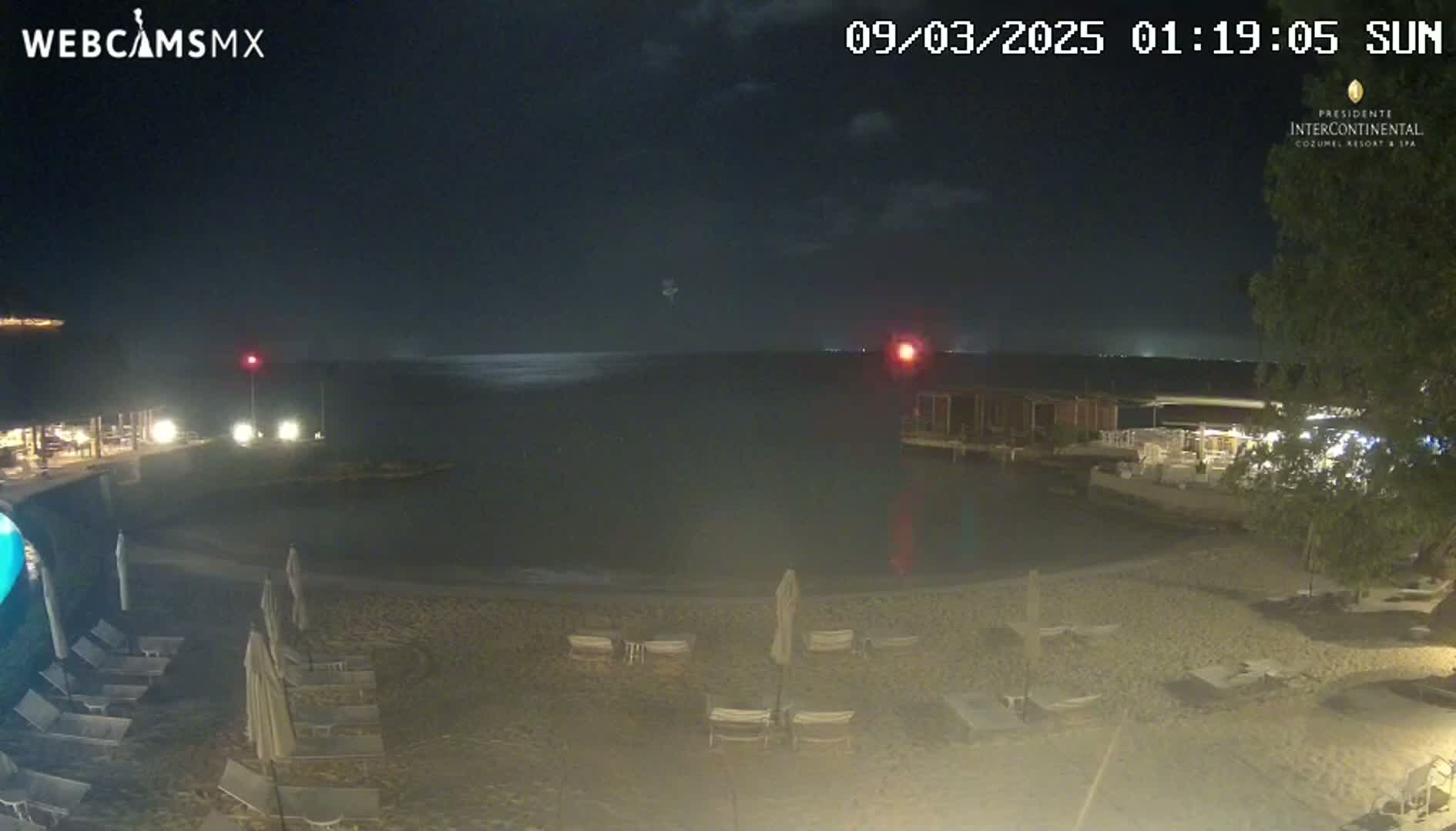 A nighttime beach scene shows a calm body of water, a sandy shore with lounge chairs and umbrellas, and low-lit buildings along the water's edge under a dark, clear sky.