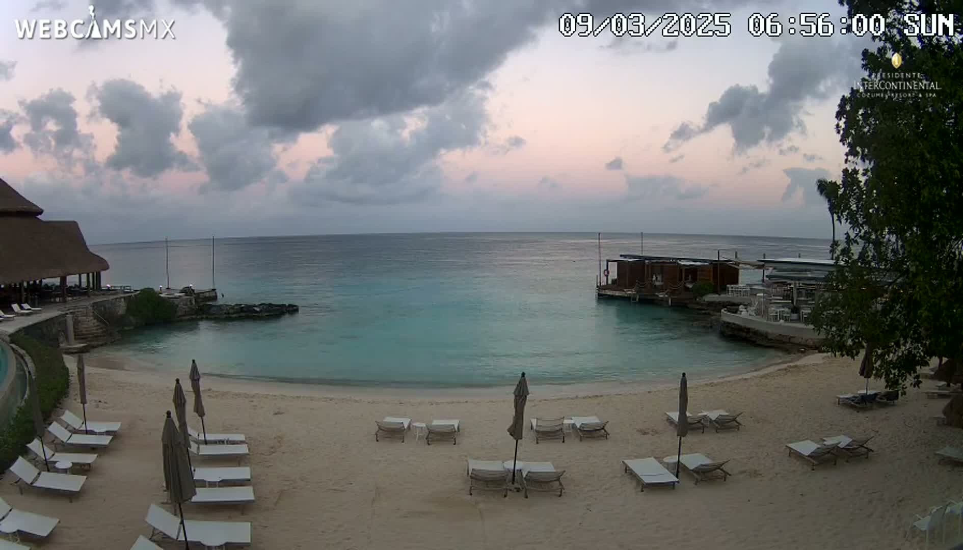 A sandy beach with lounge chairs and umbrellas overlooks calm, turquoise water under a partly cloudy sky.