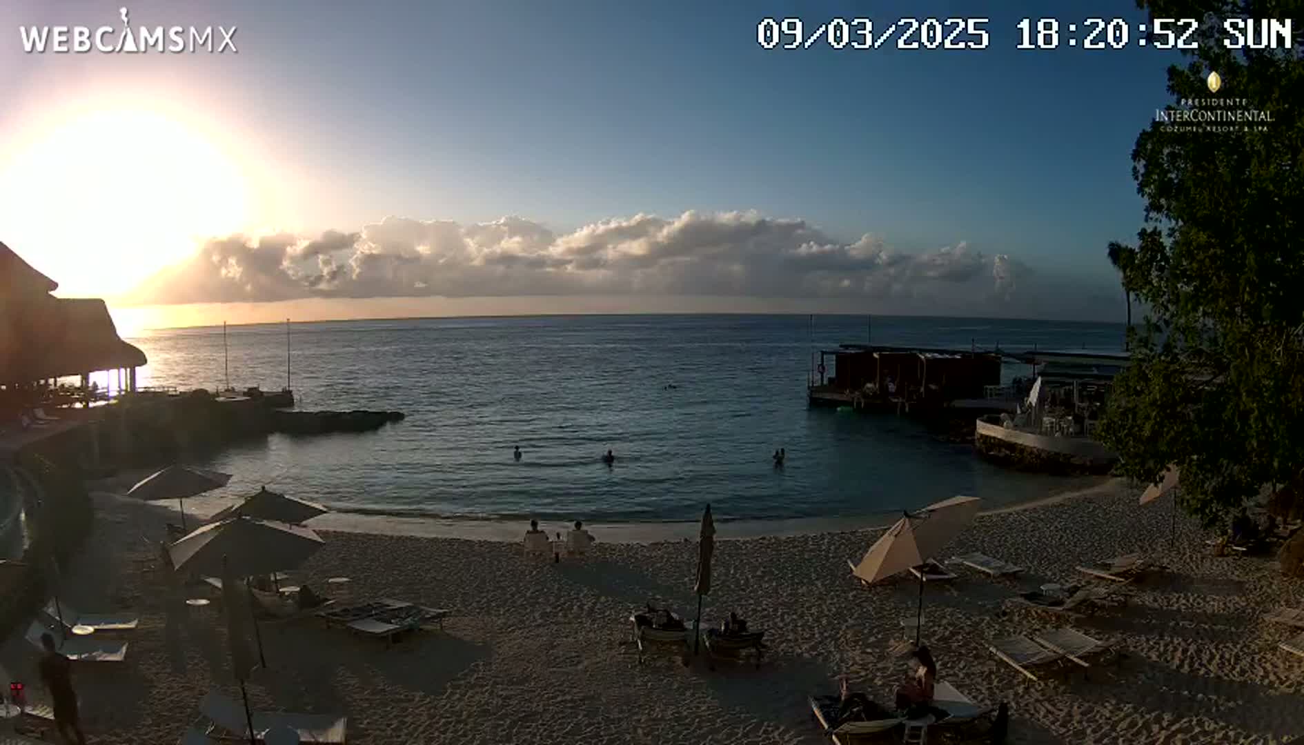 A beach scene at sunset shows calm ocean water, a sandy shore with beach umbrellas and lounge chairs, and several people swimming and relaxing.