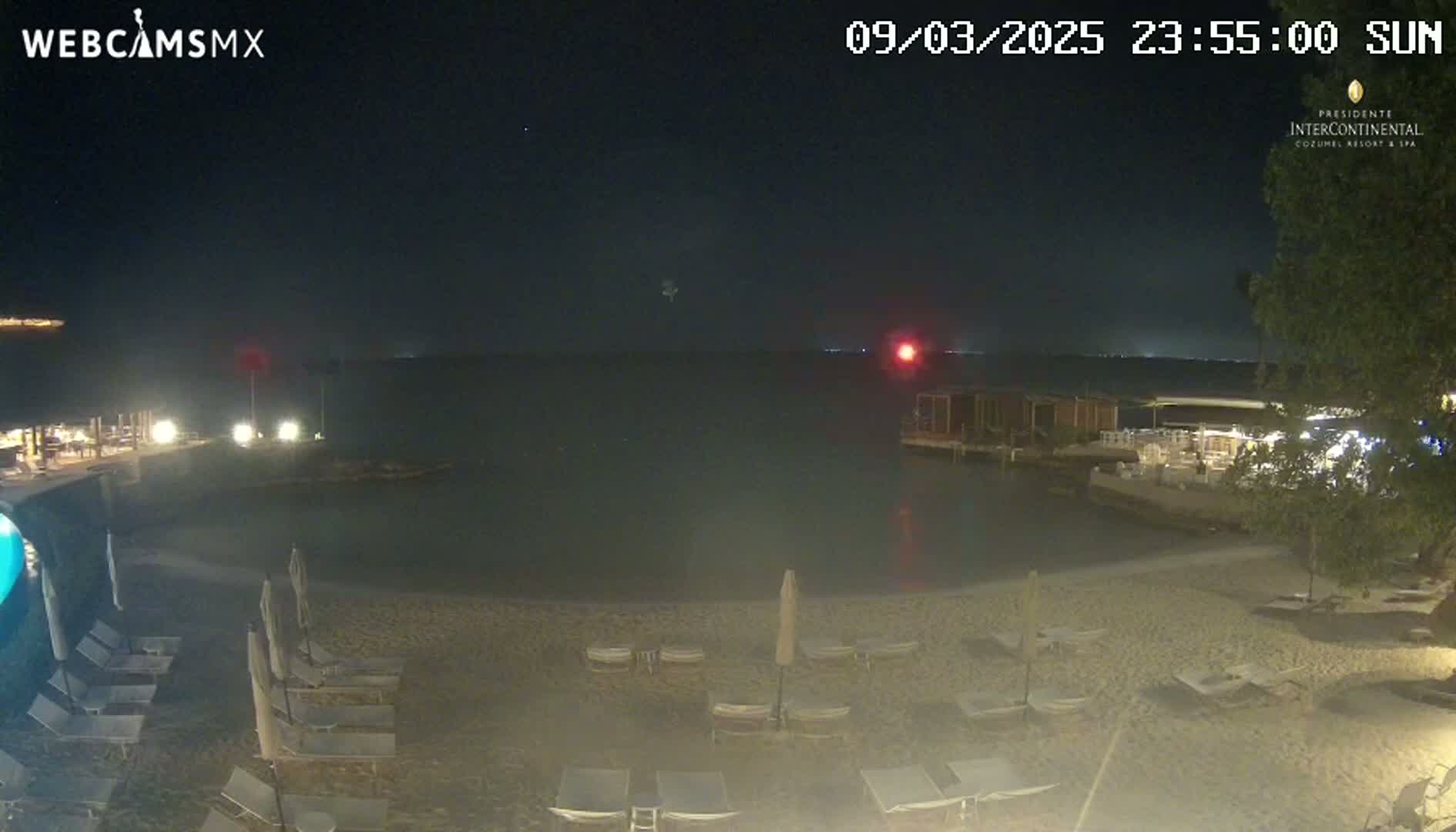 A nighttime beach scene shows calm water, dark sand, and several empty lounge chairs under closed umbrellas.