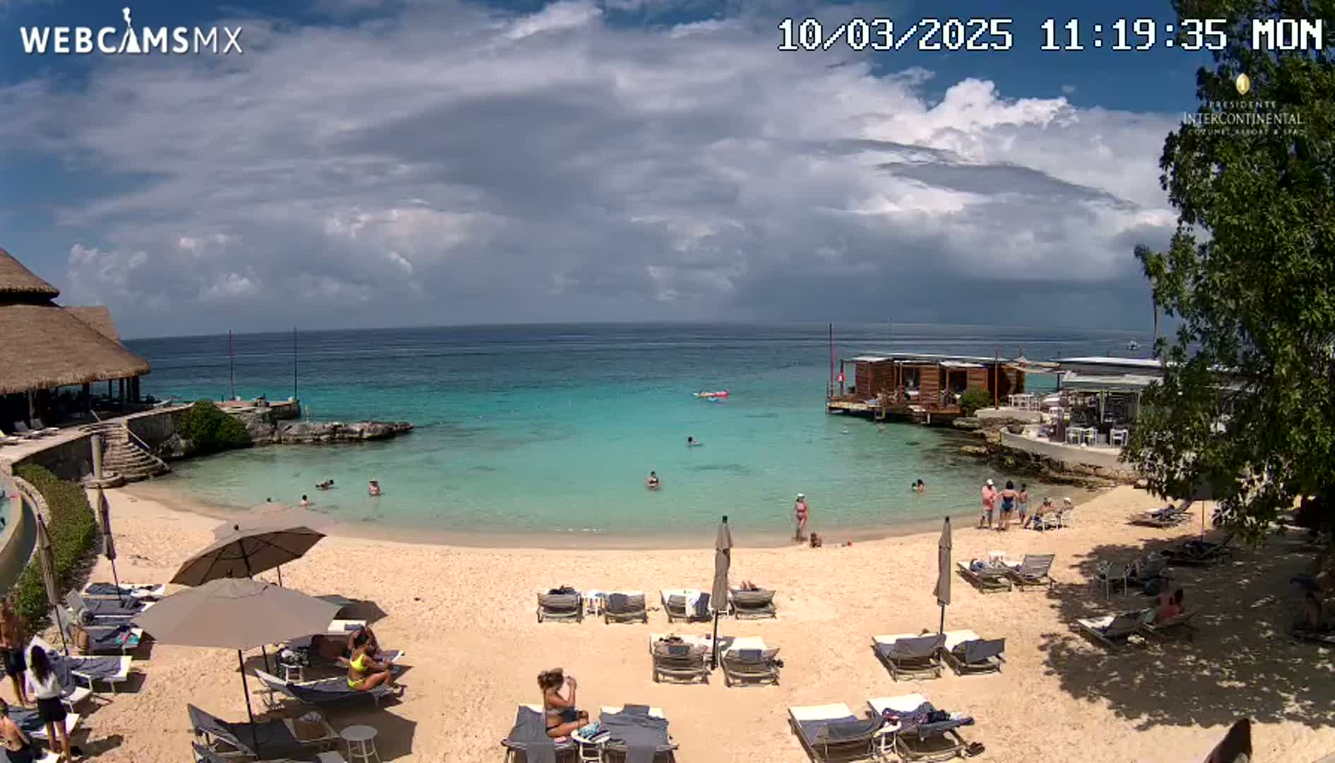 A partly cloudy day at a beach with turquoise water, a sandy shore lined with lounge chairs and umbrellas, and several people swimming and relaxing.