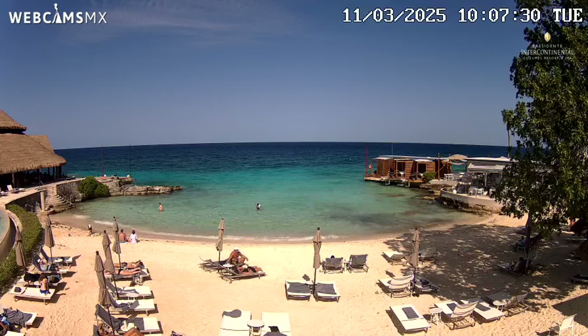A sandy beach with lounge chairs and umbrellas sits next to calm, clear turquoise water, under a sunny blue sky.
