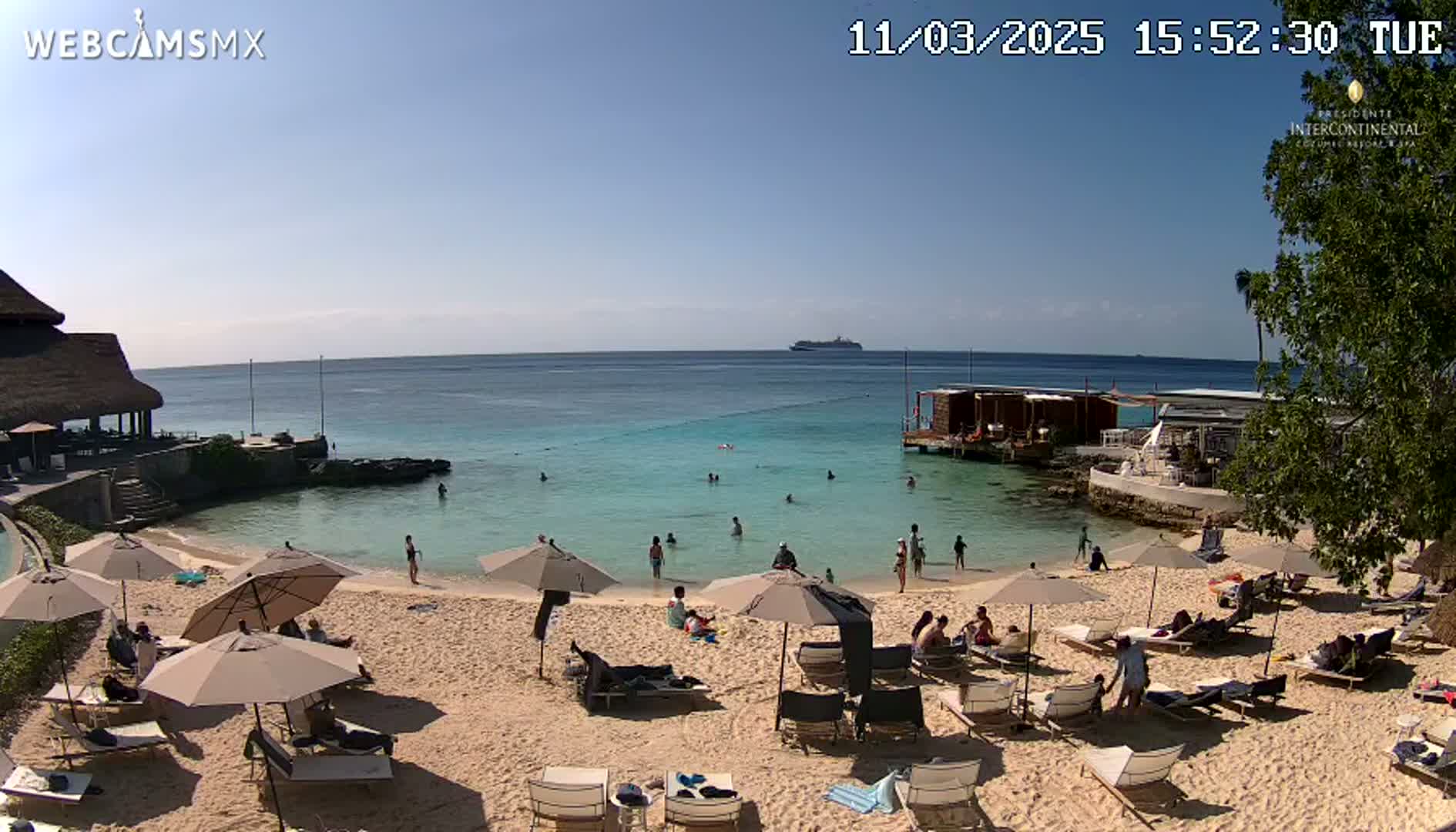 A sunny beach scene shows a calm, turquoise ocean with a cruise ship in the distance, people relaxing on lounge chairs under umbrellas, and others swimming in the shallow water near a beachfront resort.