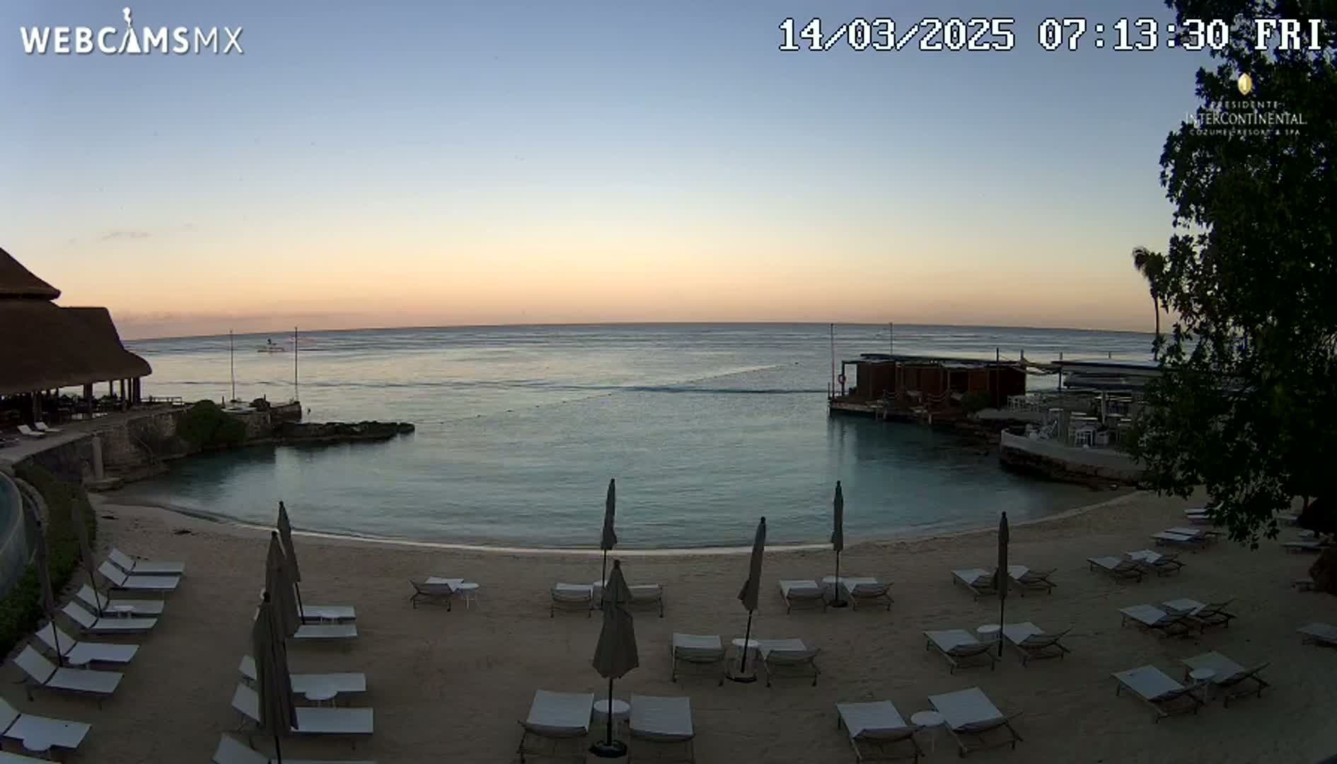 A calm, twilight ocean scene shows a sandy beach with lounge chairs and umbrellas, a building with a thatched roof, and a calm body of water.