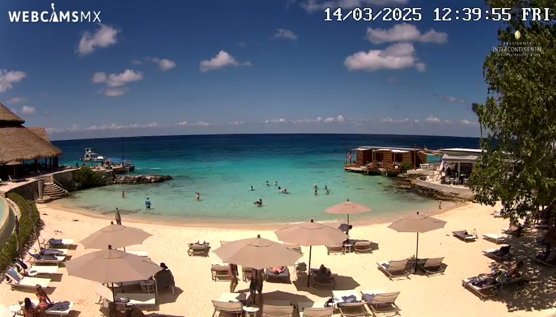 A sunny beach with calm, turquoise water, several people swimming and lounging under umbrellas, and buildings along the shoreline.