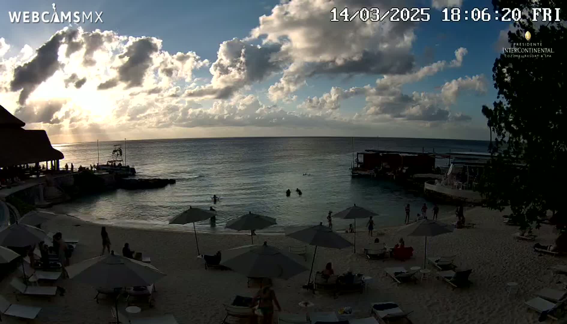 A beach scene at sunset shows people relaxing under umbrellas on a sandy beach near calm ocean water, with partly cloudy skies.