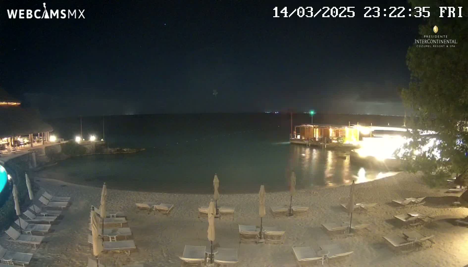 A nighttime beach scene shows a sandy shoreline with lounge chairs and umbrellas under a clear, dark sky, and a calm body of water with a structure extending into it.