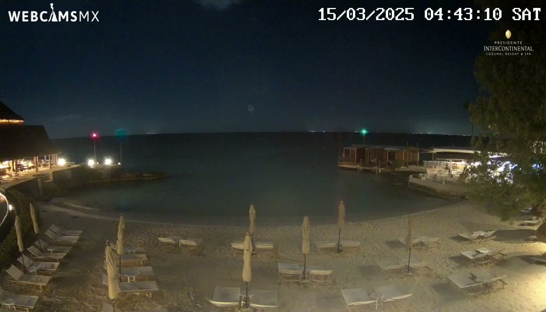 A nighttime view of a beach with lounge chairs and umbrellas under a clear, dark sky, showing calm water and a resort in the background.