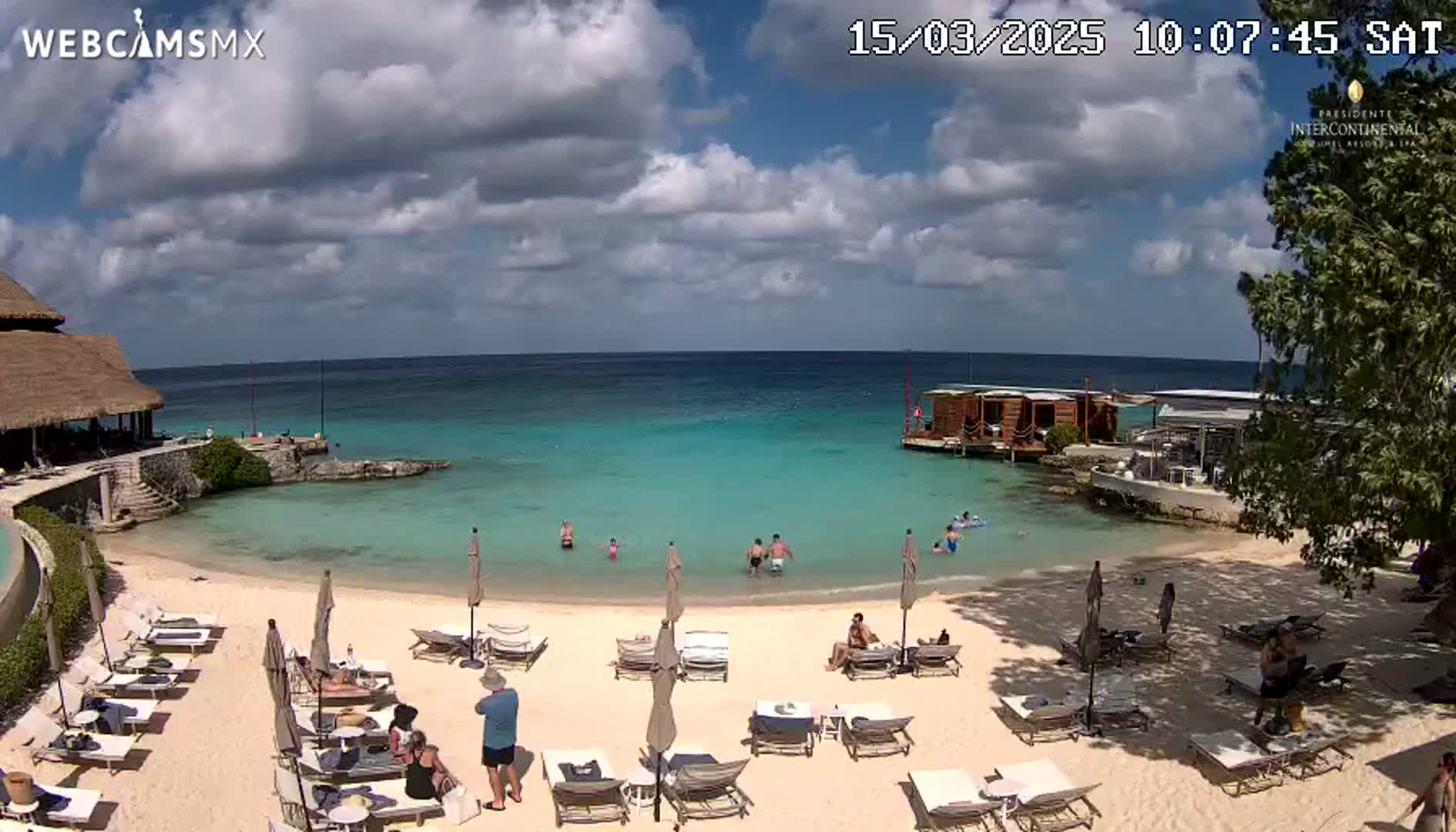 A partly cloudy sunny day at a beach resort shows a sandy beach with lounge chairs and umbrellas, turquoise water with people swimming, and buildings including a thatched-roof structure and a wooden overwater structure.