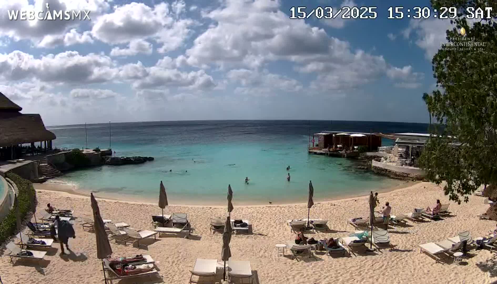 A partly cloudy day at a beach with calm, turquoise water, several people swimming and lounging on chairs under umbrellas on a sandy beach near some buildings.