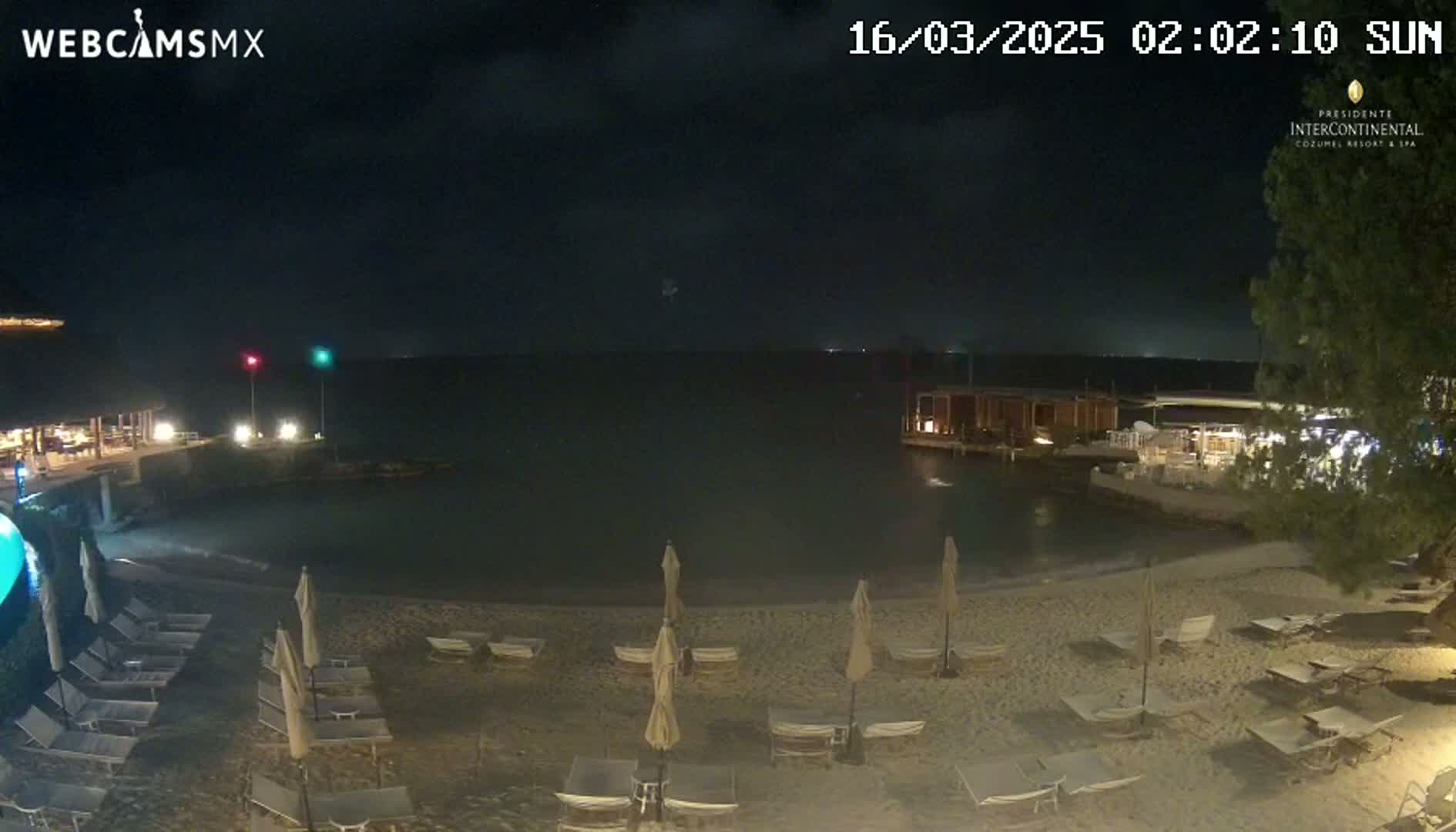 A nighttime beach scene shows a sandy shoreline with lounge chairs and umbrellas under a dark sky over calm water.