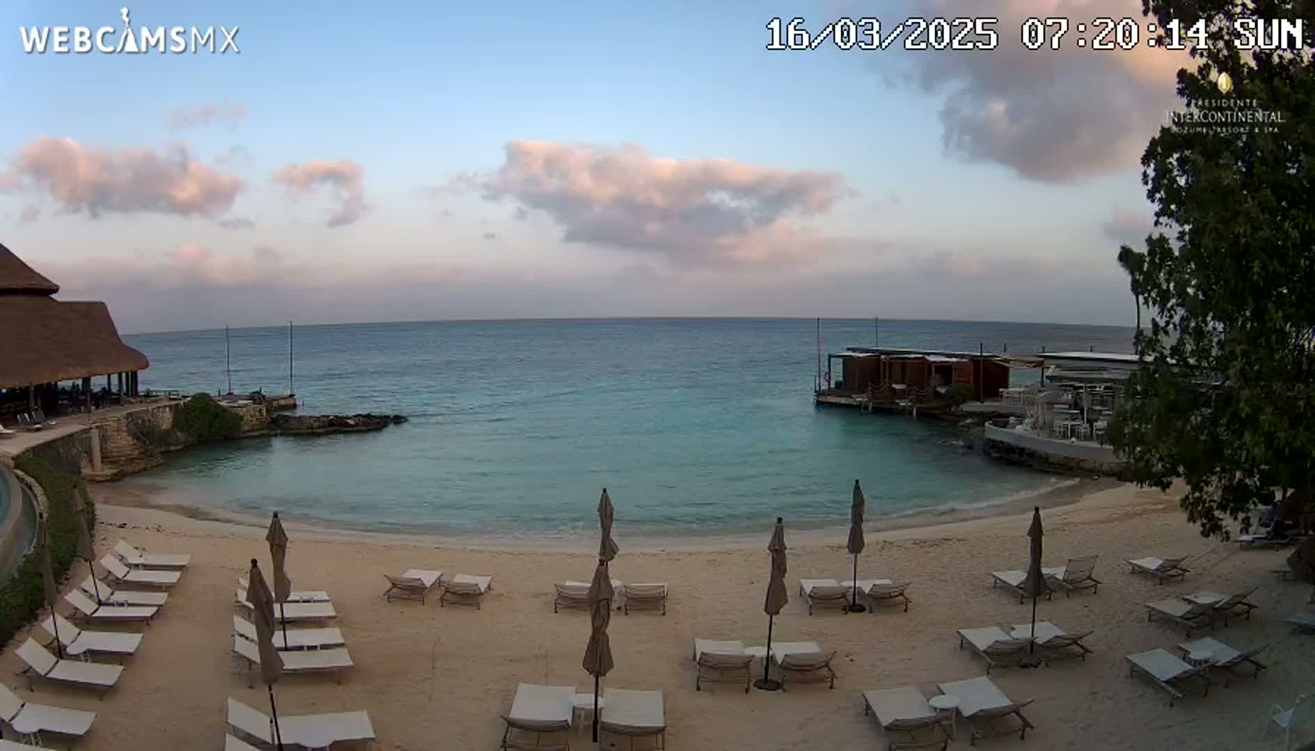 A tranquil beach scene at sunrise features calm, clear water, numerous empty lounge chairs under umbrellas, and a building with a thatched roof.