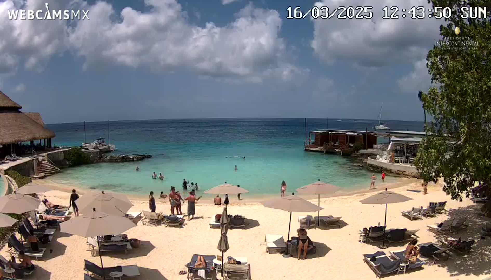 A sunny day on a beach with clear turquoise water, several people swimming and sunbathing, and lounge chairs and umbrellas set up on the sand.