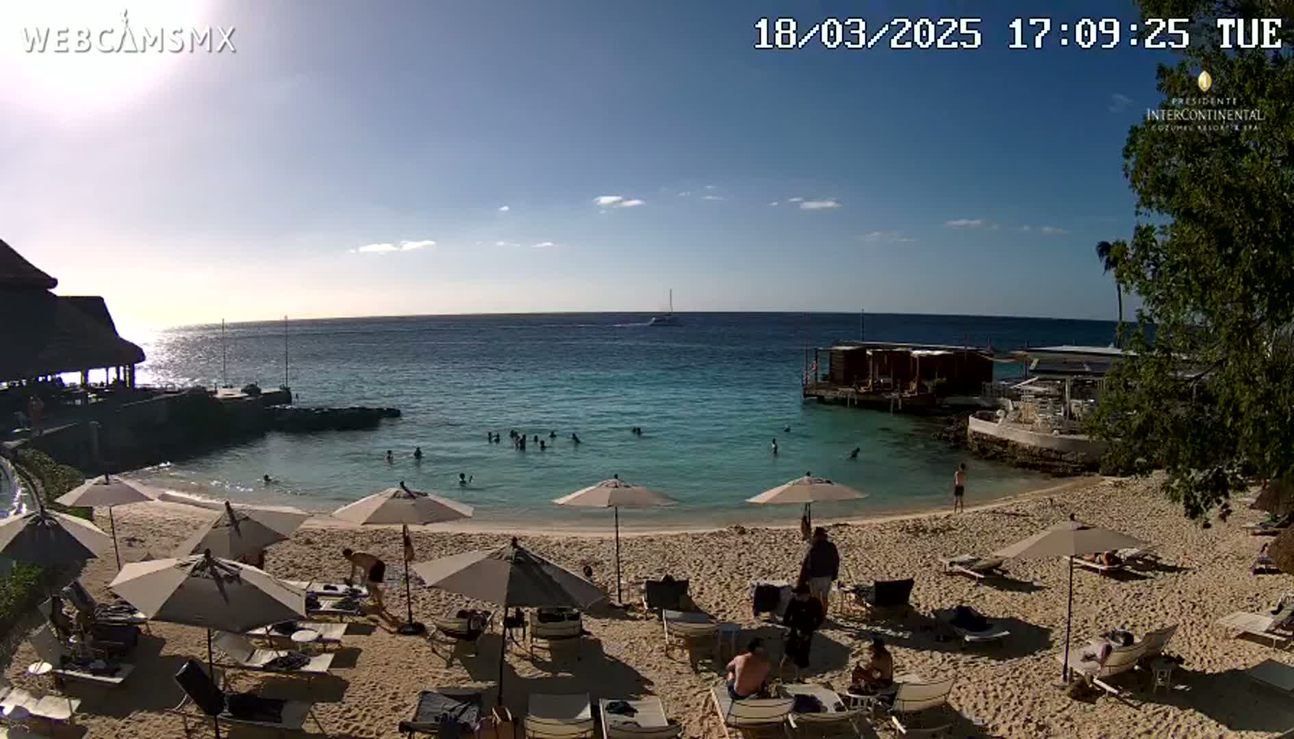 A sandy beach with calm, clear turquoise water, numerous beach umbrellas and lounge chairs, and several people swimming and relaxing under a mostly sunny sky.