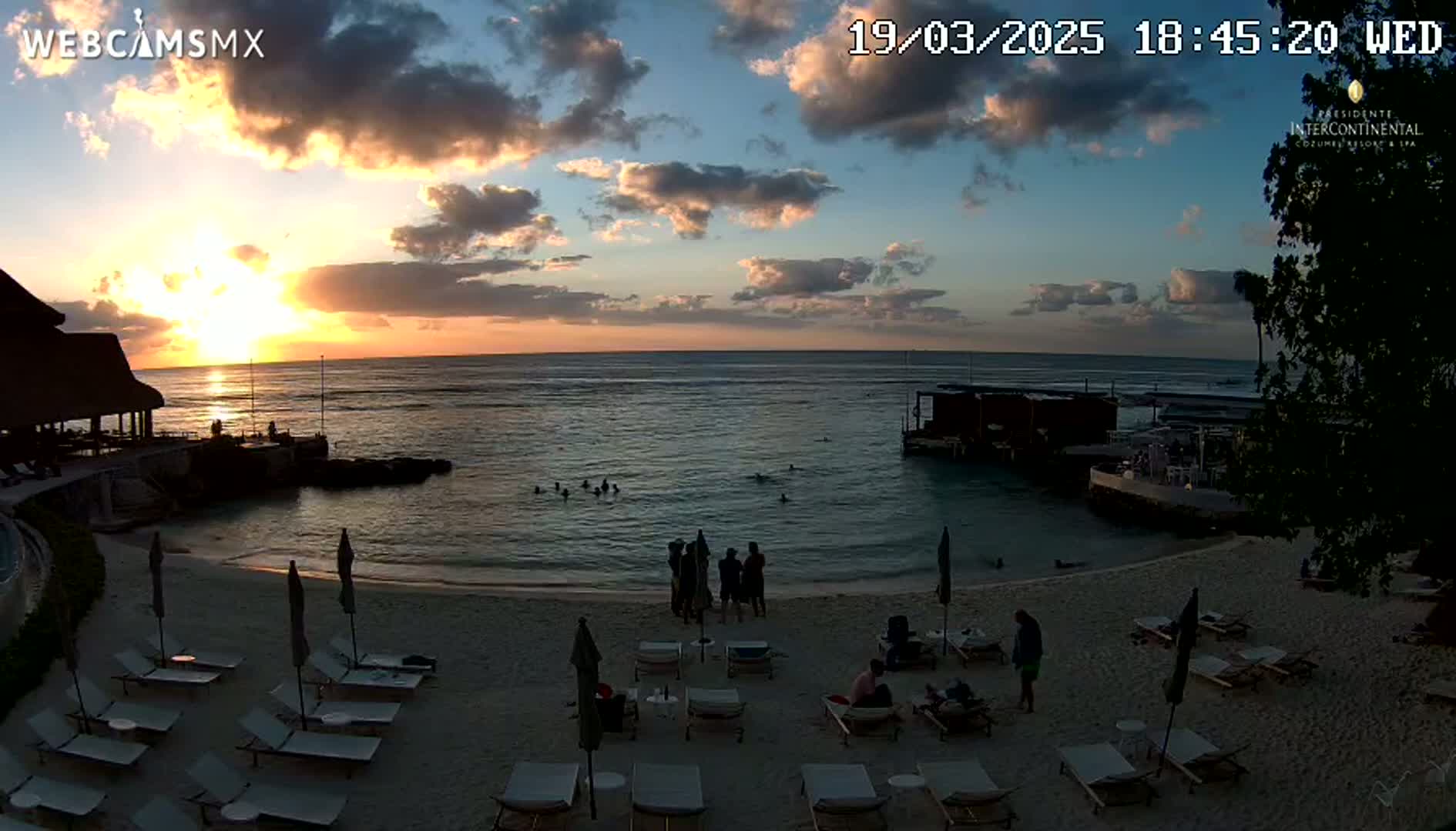 A beach scene at sunset shows people relaxing on lounge chairs near calm ocean water with a few people swimming, under a partly cloudy sky.