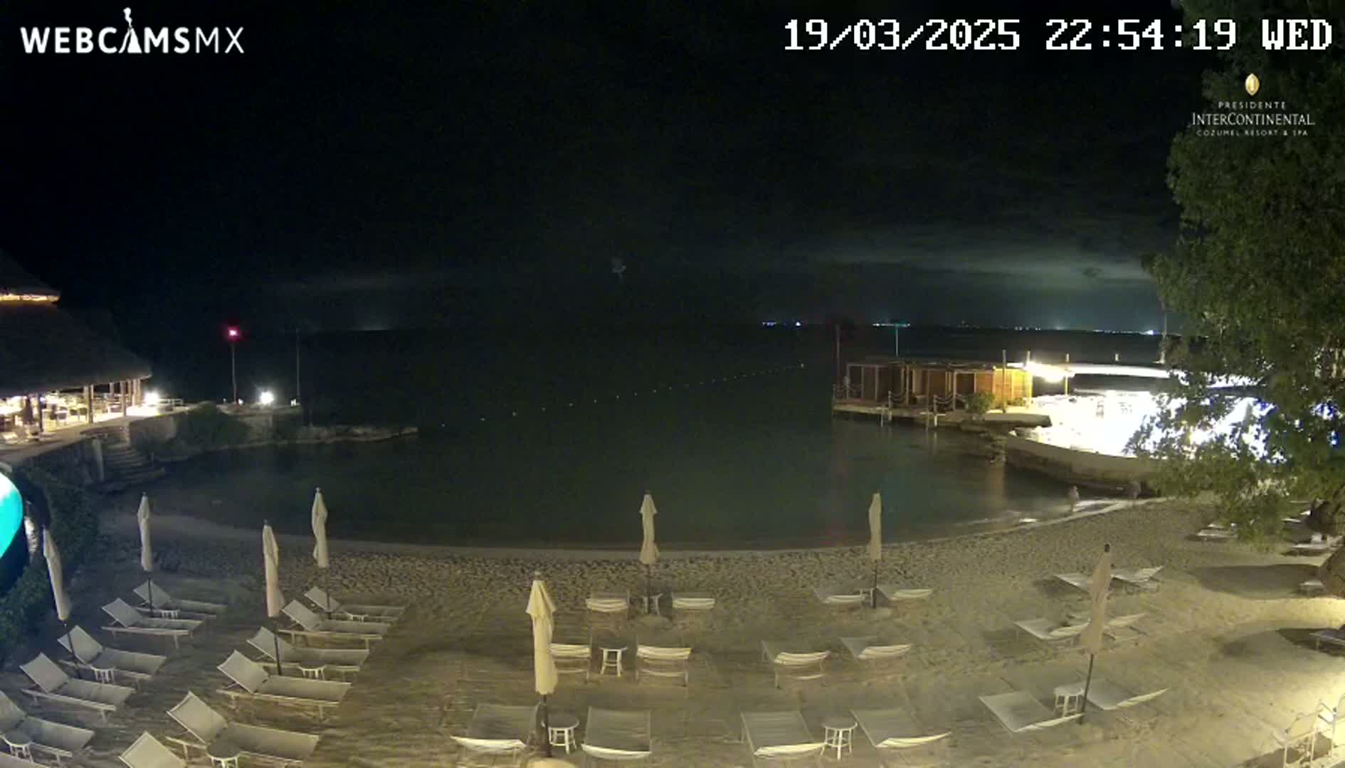 A nighttime beach scene shows a sandy shore lined with lounge chairs and umbrellas under a partly cloudy sky, next to calm water with a building visible across the bay.