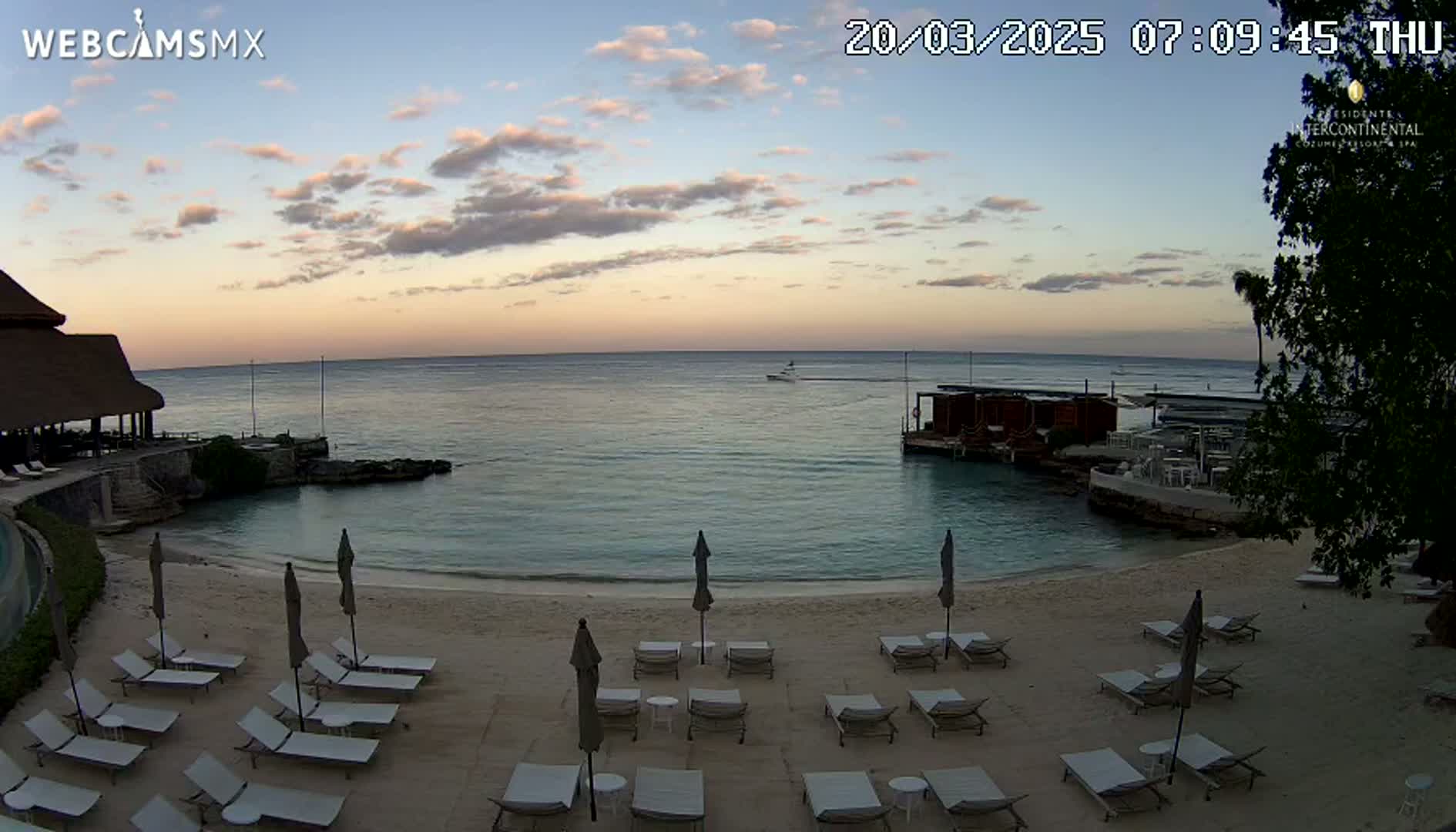 A sandy beach with lounge chairs and umbrellas sits beside calm, twilight ocean waters under a partly cloudy sky.