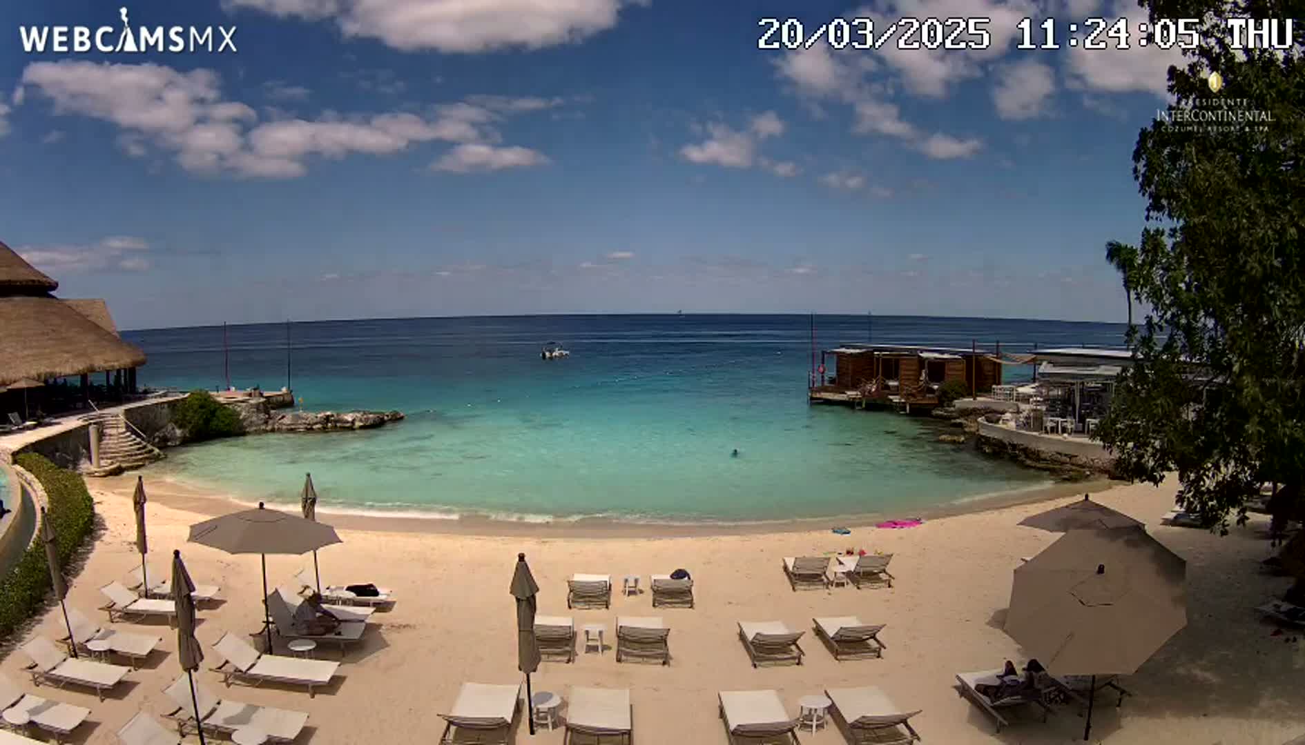 A sunny day at a beach with calm, turquoise water, a sandy shore lined with lounge chairs and umbrellas, and a building with a thatched roof in the background.