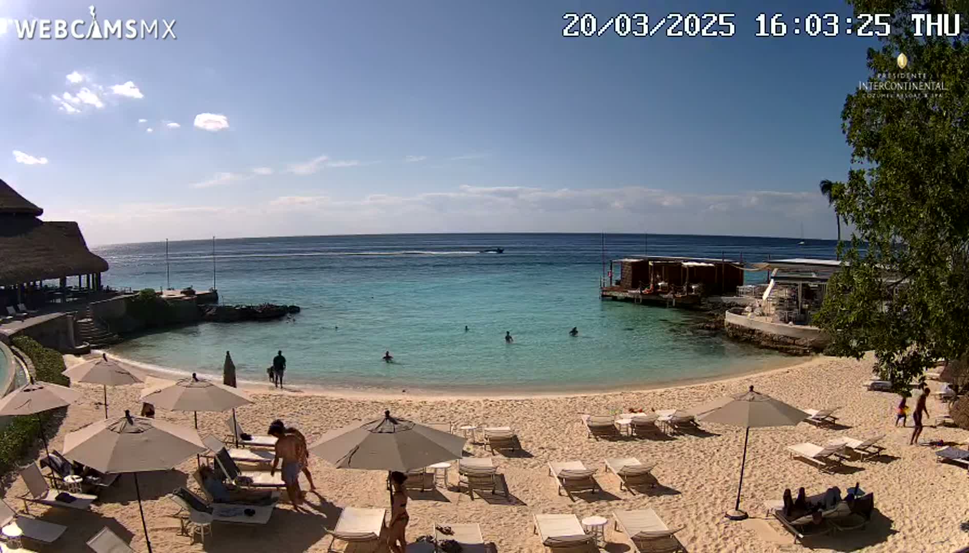 A sunny beach scene shows a calm, light-blue ocean with several people swimming, and a sandy beach with lounge chairs and umbrellas, some people relaxing on the beach, and resort buildings nearby.