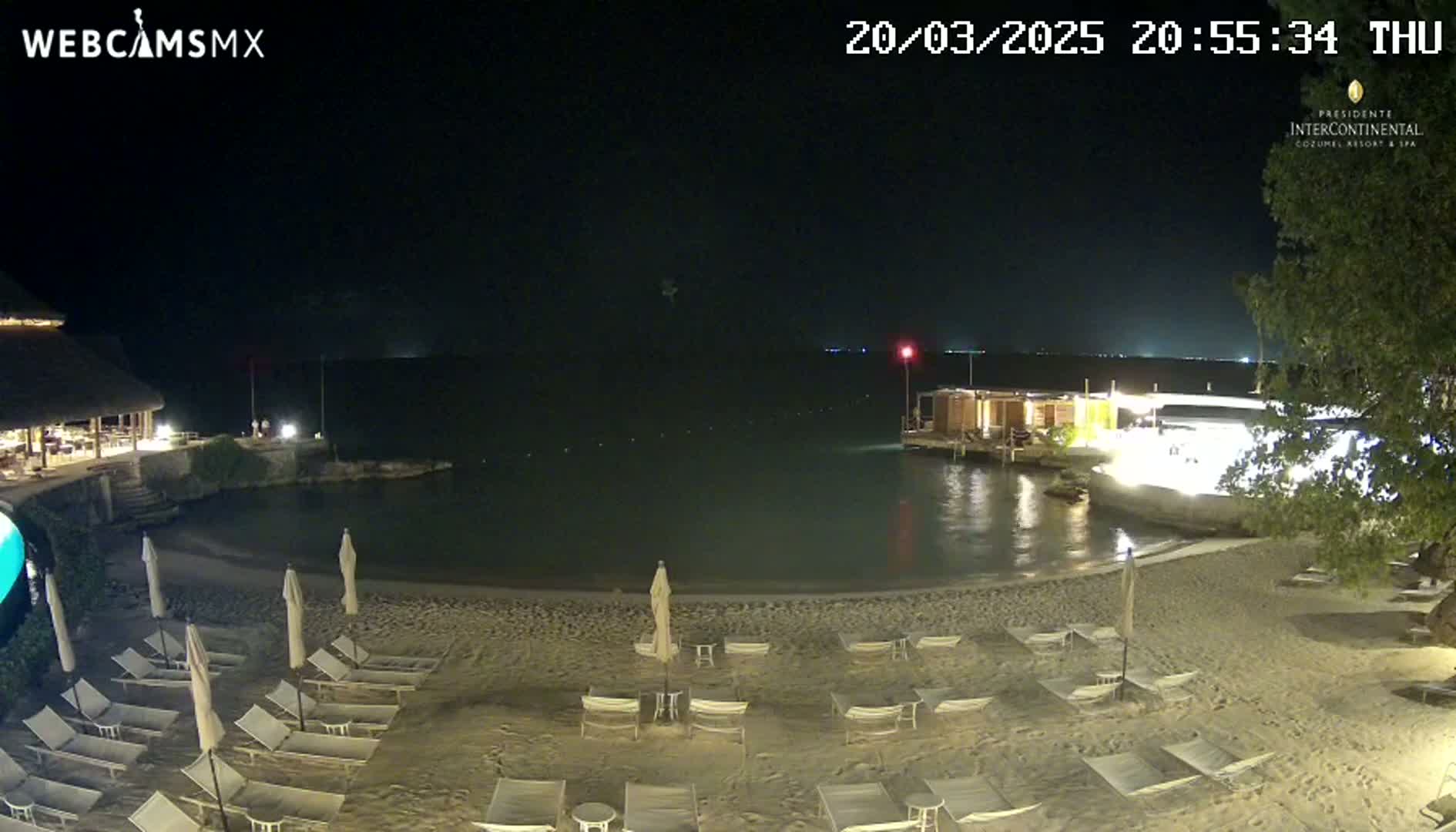 A nighttime beach scene shows a calm body of water, a sandy shoreline with numerous empty lounge chairs and umbrellas under soft lighting, and a building partially visible in the background.