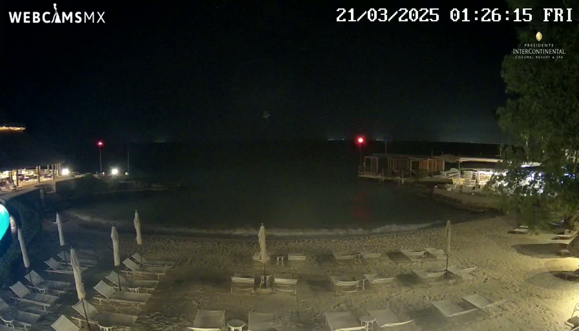 A nighttime view of a sandy beach with lounge chairs and umbrellas, a calm body of water, and some buildings in the background under a clear, dark sky.