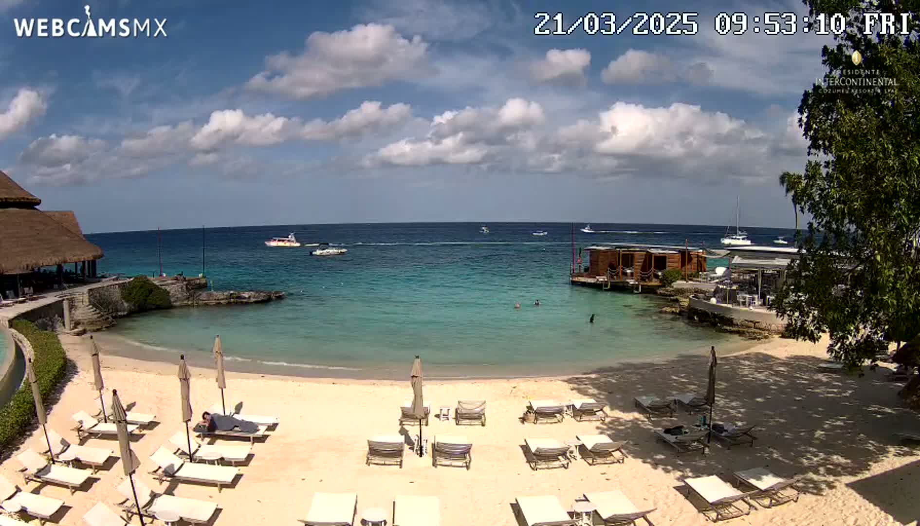 A sandy beach with lounge chairs and umbrellas overlooks calm, turquoise water with several boats visible in the distance under a partly cloudy sky.
