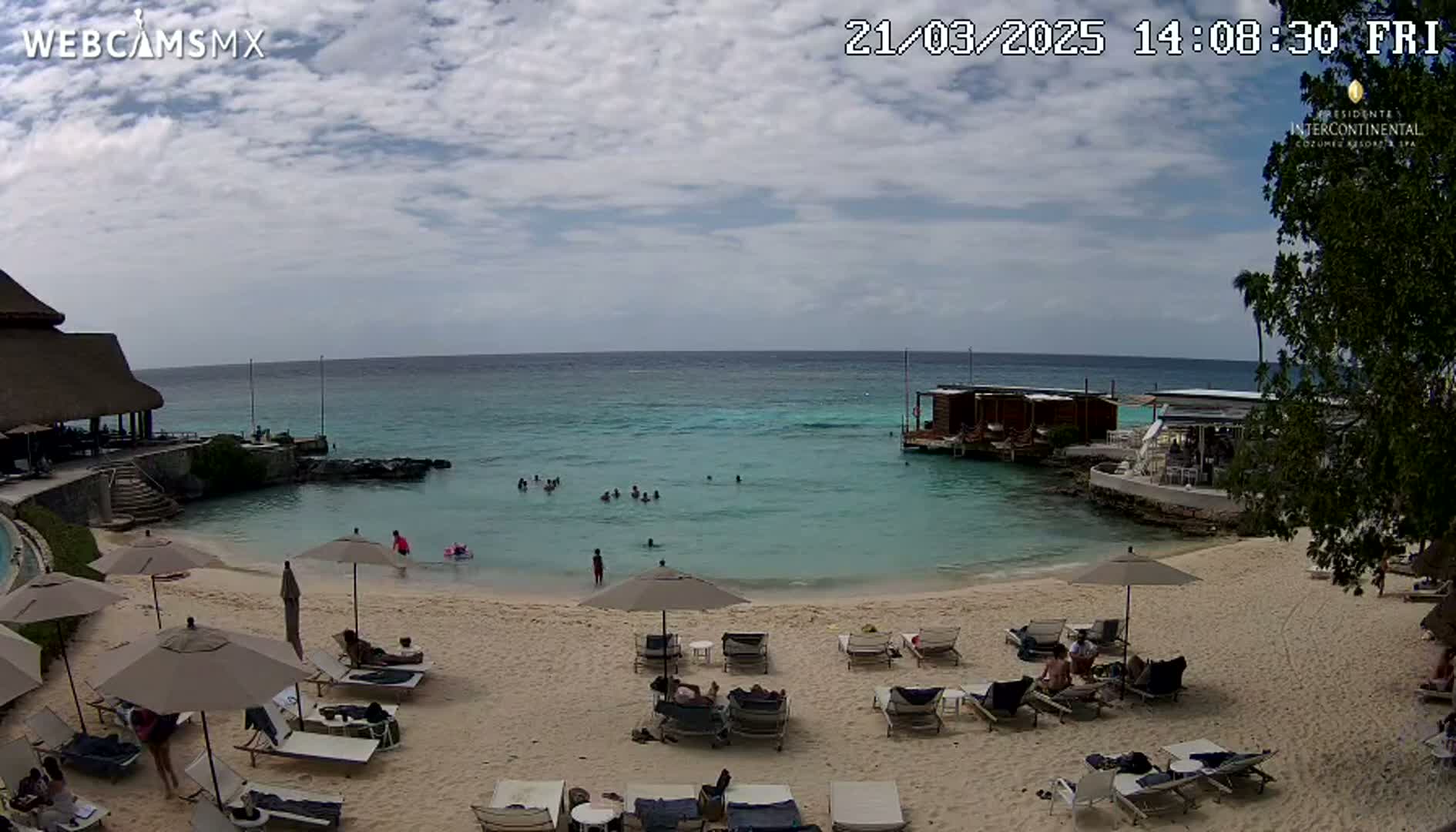 A sandy beach with lounge chairs and umbrellas under a partly cloudy sky, overlooks a calm, turquoise ocean where people are swimming near a small, wooden overwater structure.