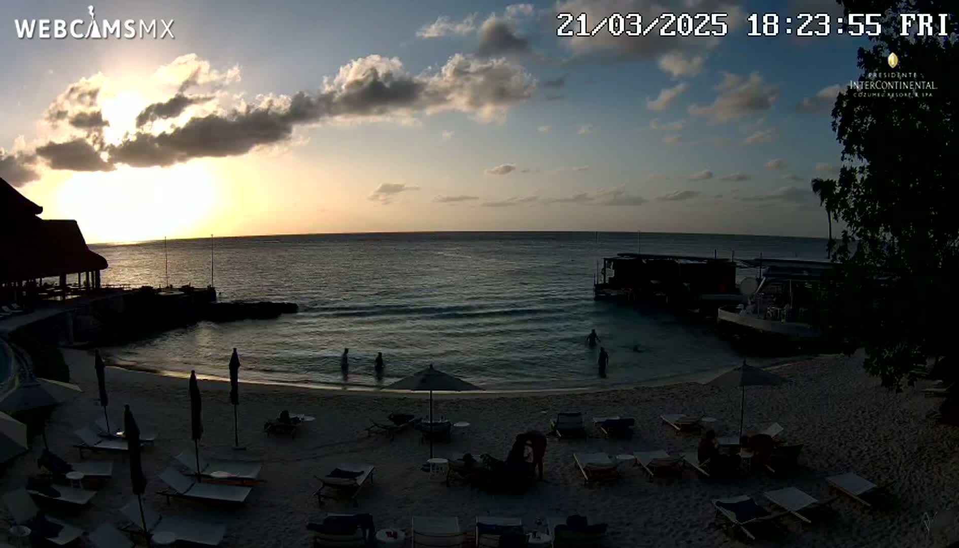 A beach scene at sunset shows a calm ocean, a sandy shore with lounge chairs and umbrellas, and a few people in the water.