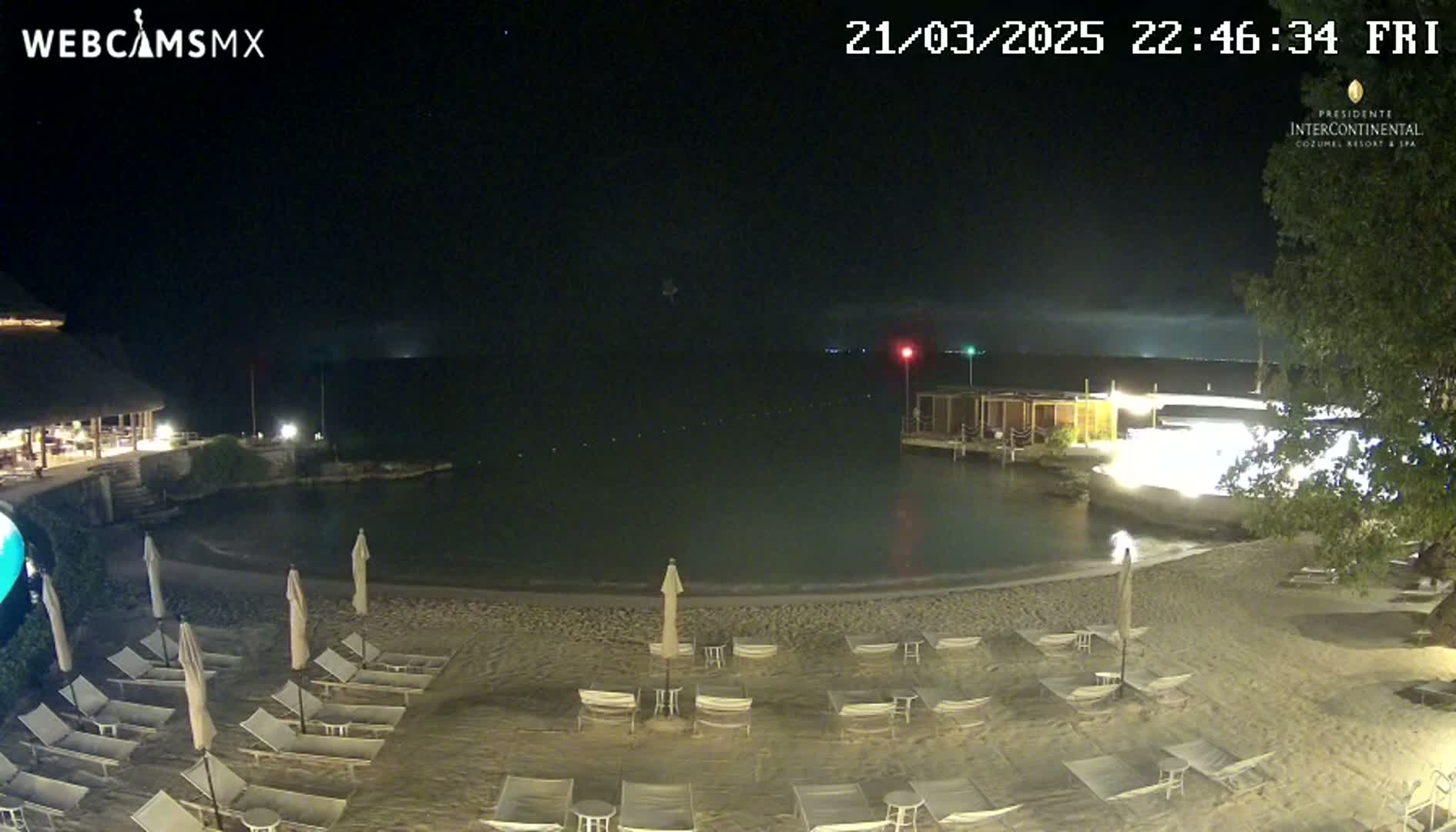 A nighttime beach scene shows a calm body of water, a sandy shore with empty lounge chairs under umbrellas, and a building with lights in the background under a clear, starry sky.