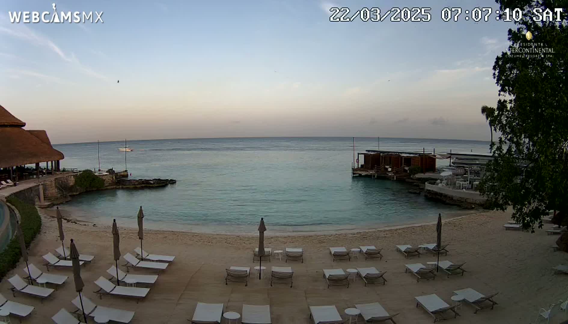 A sandy beach with lounge chairs and umbrellas sits beside calm, clear turquoise water, next to a resort building under a mostly clear sky at dawn.