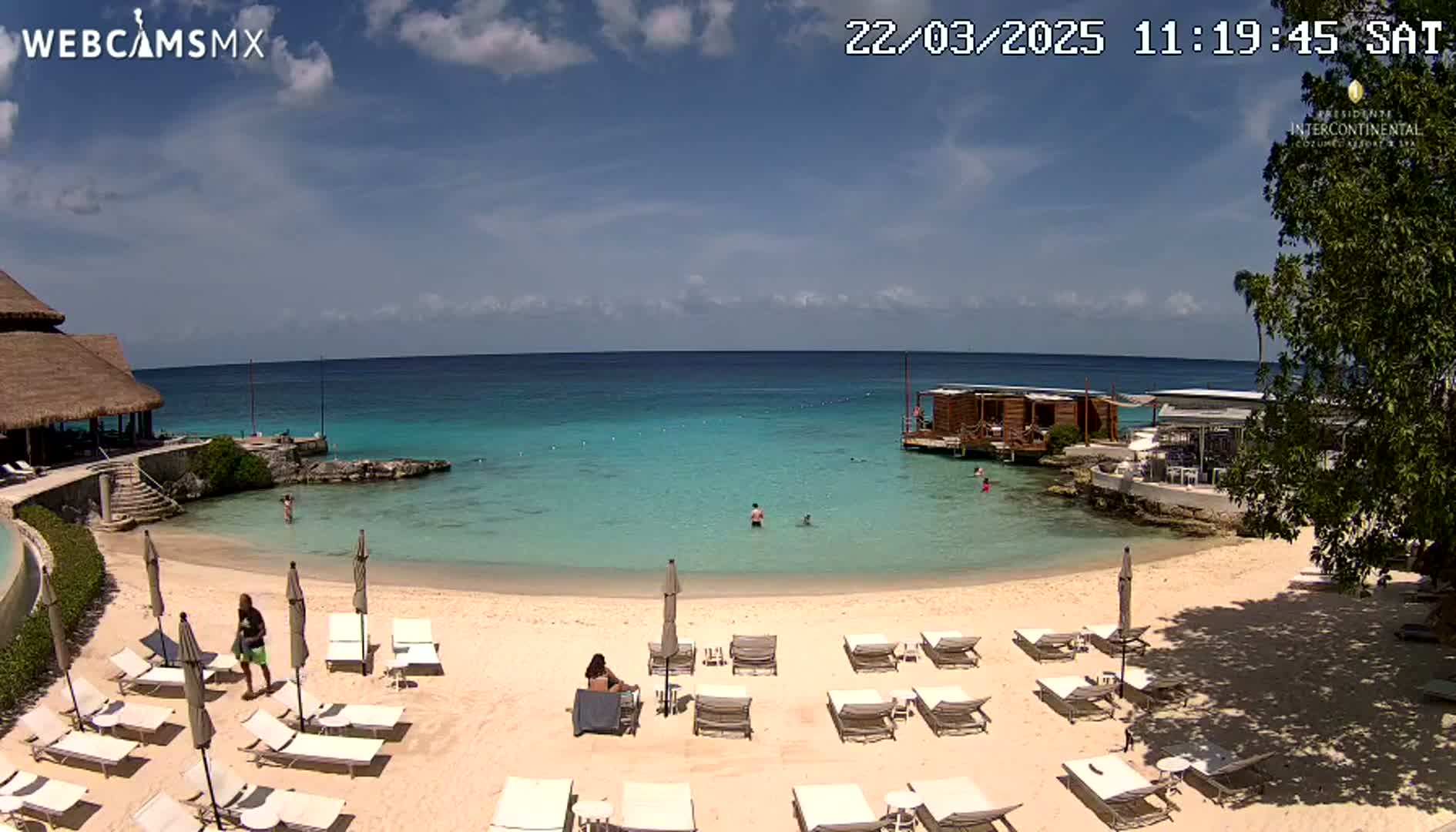 A beach with lounge chairs and umbrellas under a partly cloudy sky, overlooks calm turquoise water and a small overwater structure.