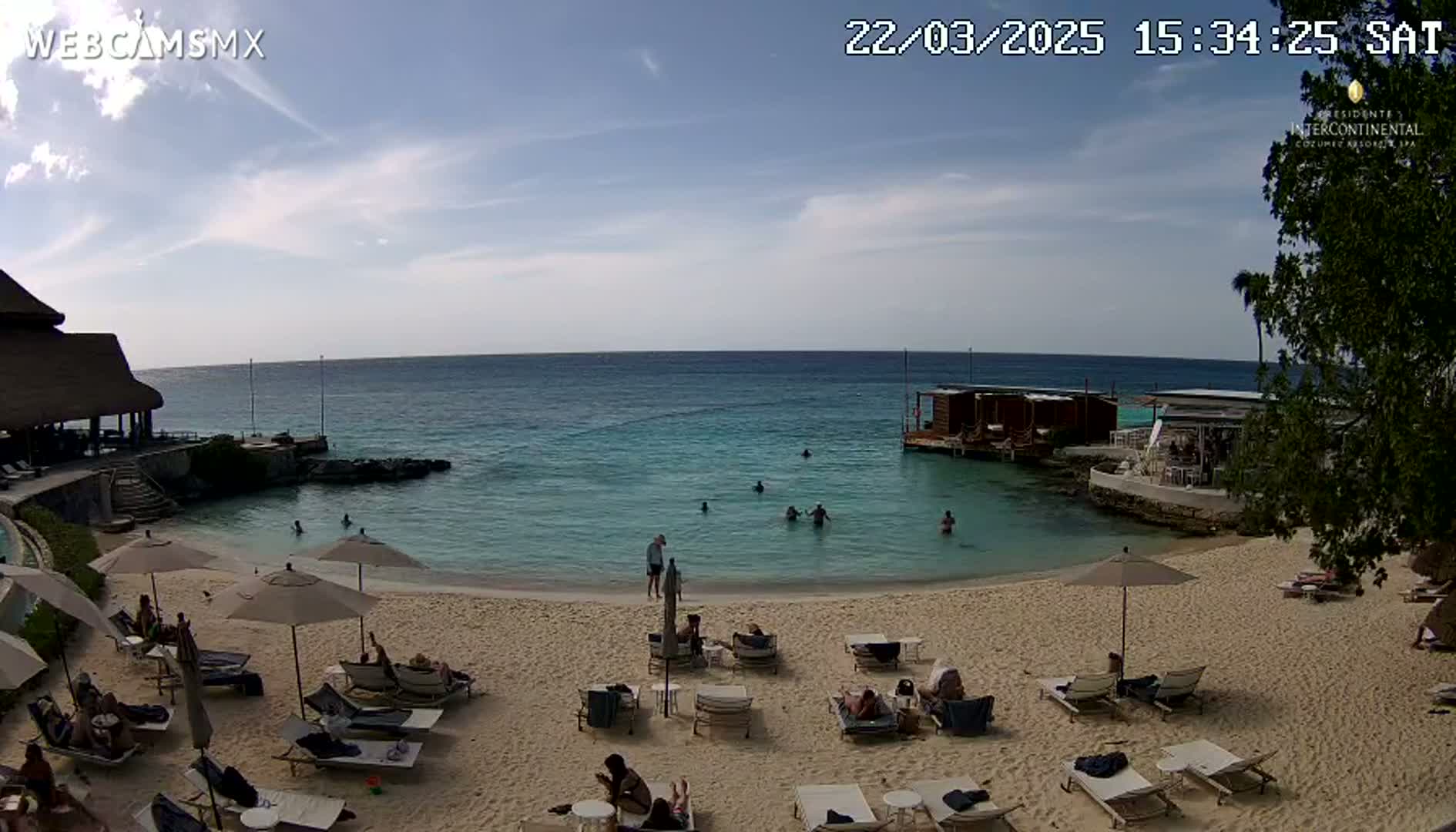 A sandy beach with lounge chairs and umbrellas under a mostly sunny sky, featuring calm, clear turquoise water with several people swimming and relaxing.