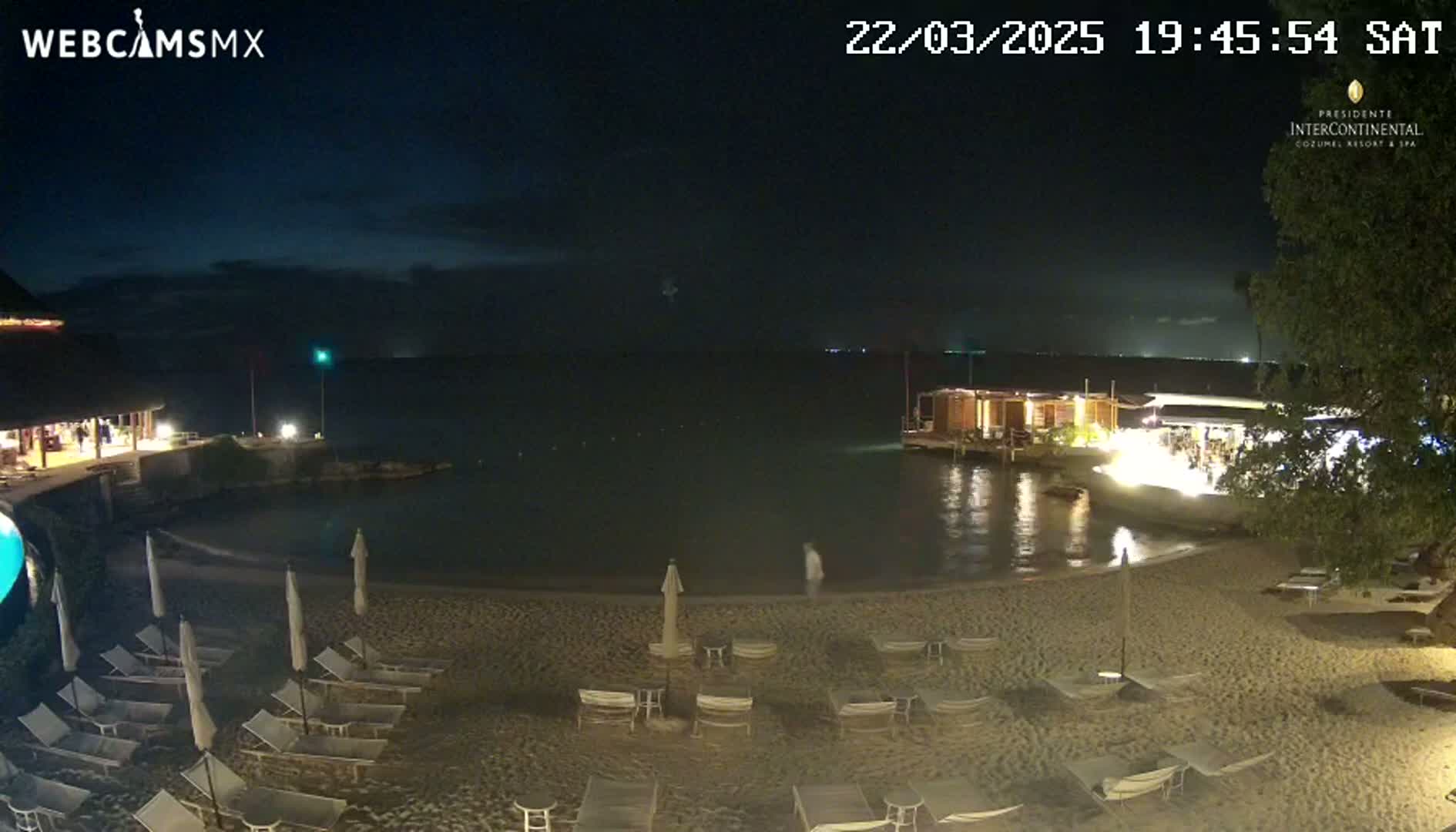 A nighttime beach scene shows a calm body of water, a sandy beach with lounge chairs, and low-light buildings along the shore under a clear, dark sky.