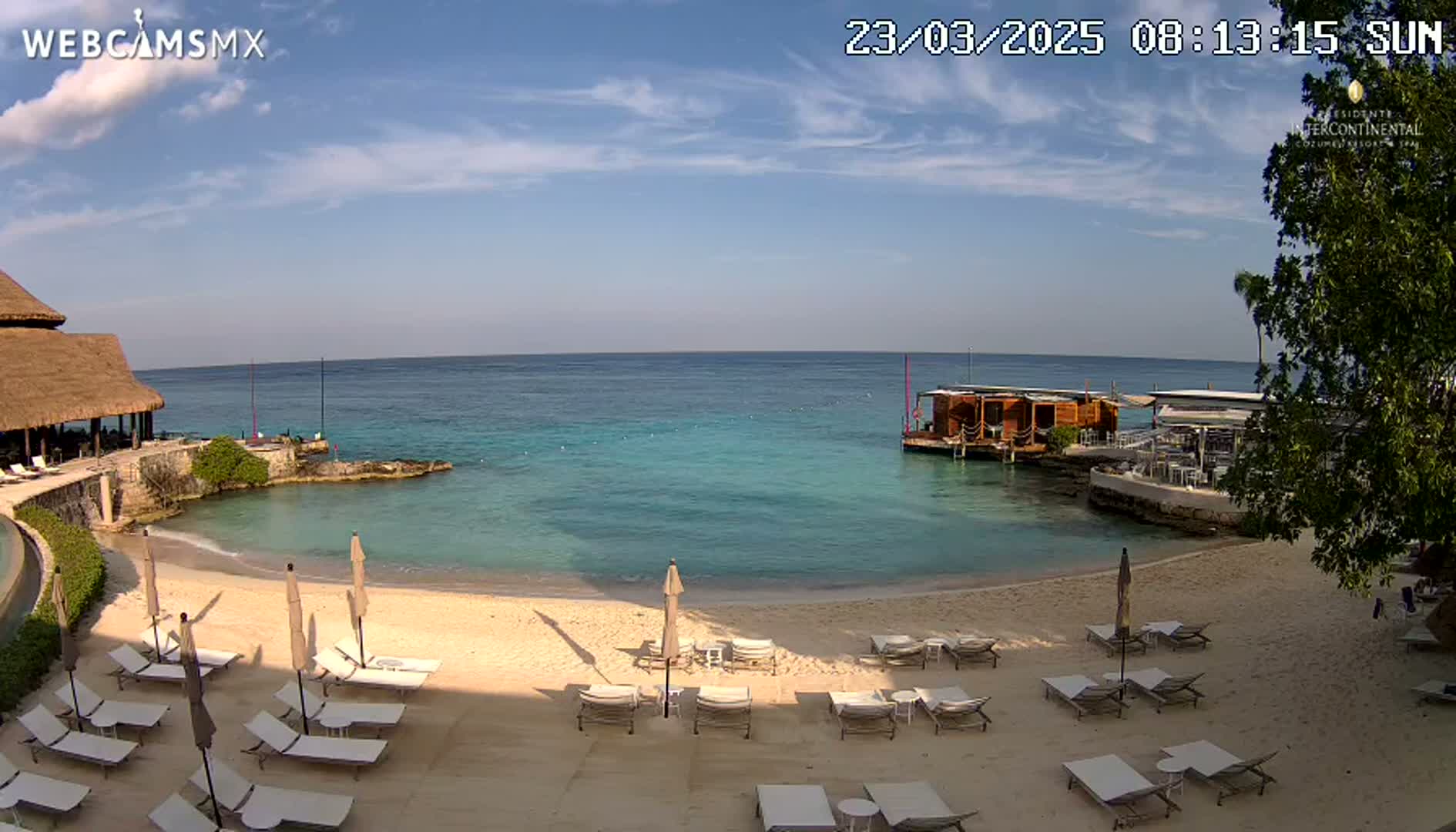A sandy beach with lounge chairs and umbrellas is situated next to a calm, turquoise ocean, under a mostly sunny sky.
