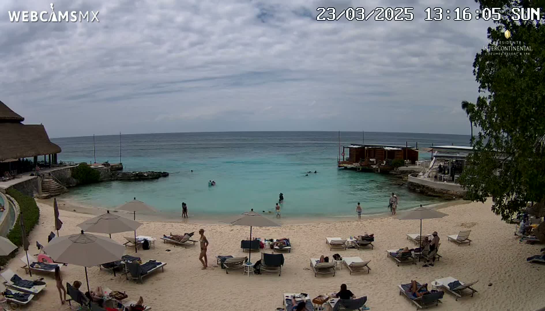 A sandy beach with turquoise water, several people relaxing under umbrellas, and a building on the water under a partly cloudy sky.