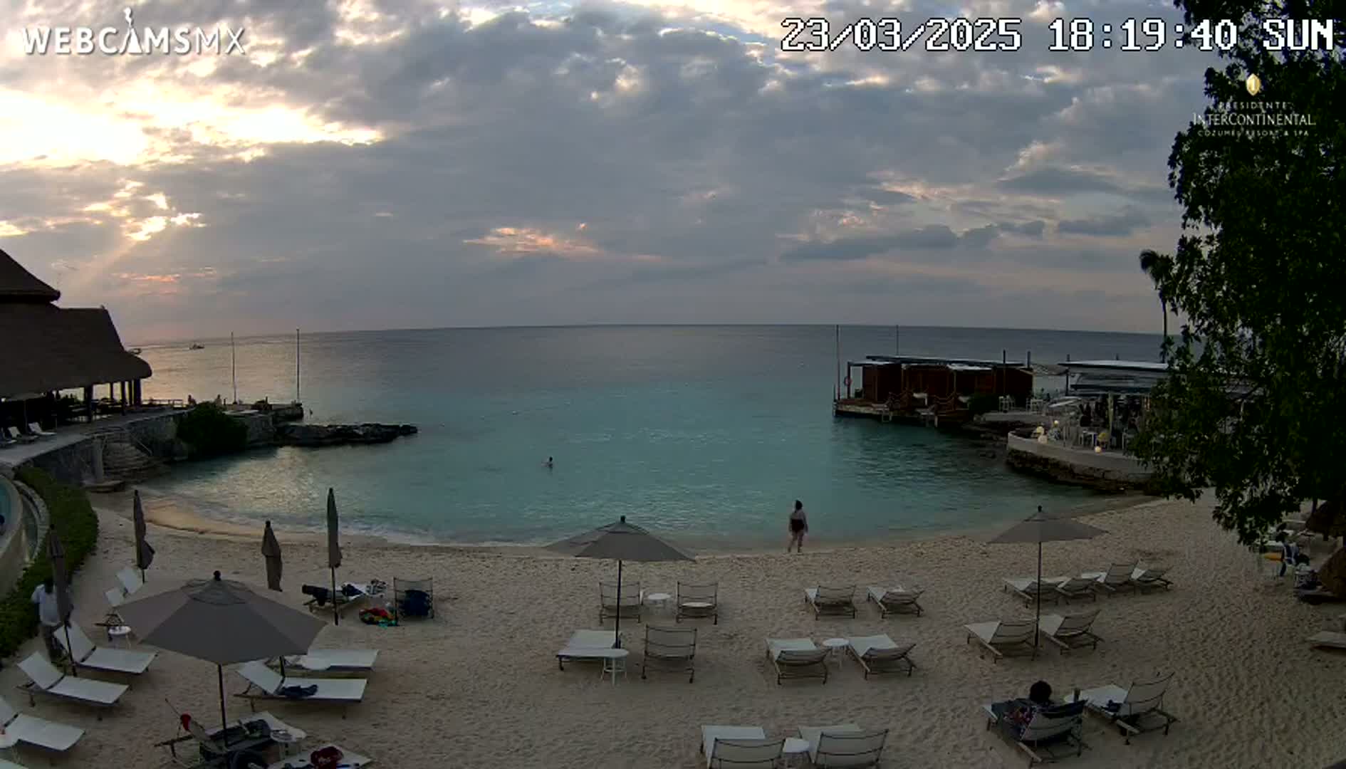 A sandy beach with lounge chairs and umbrellas sits beside calm turquoise water under a partly cloudy sunset sky.