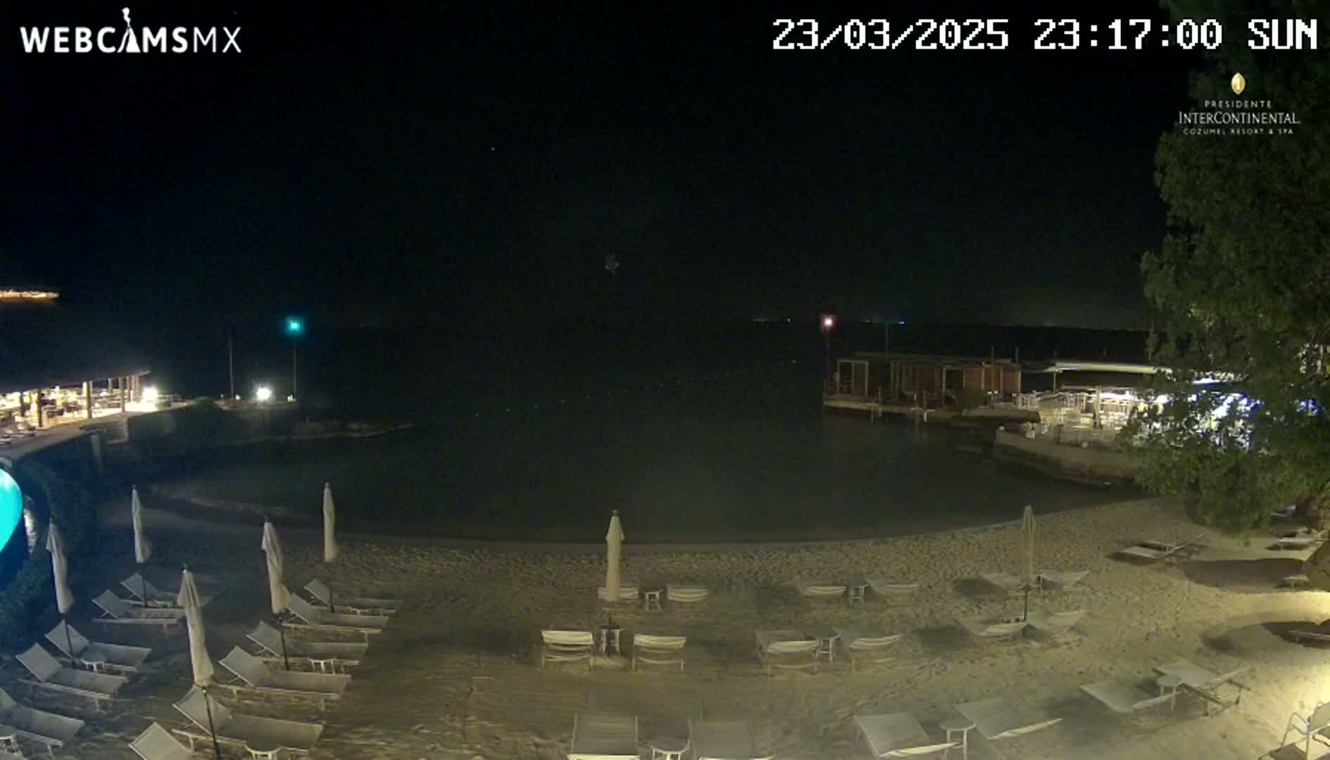 A nighttime beach scene shows empty lounge chairs under beach umbrellas surrounding a calm body of water with some buildings visible in the background under a clear, dark sky.