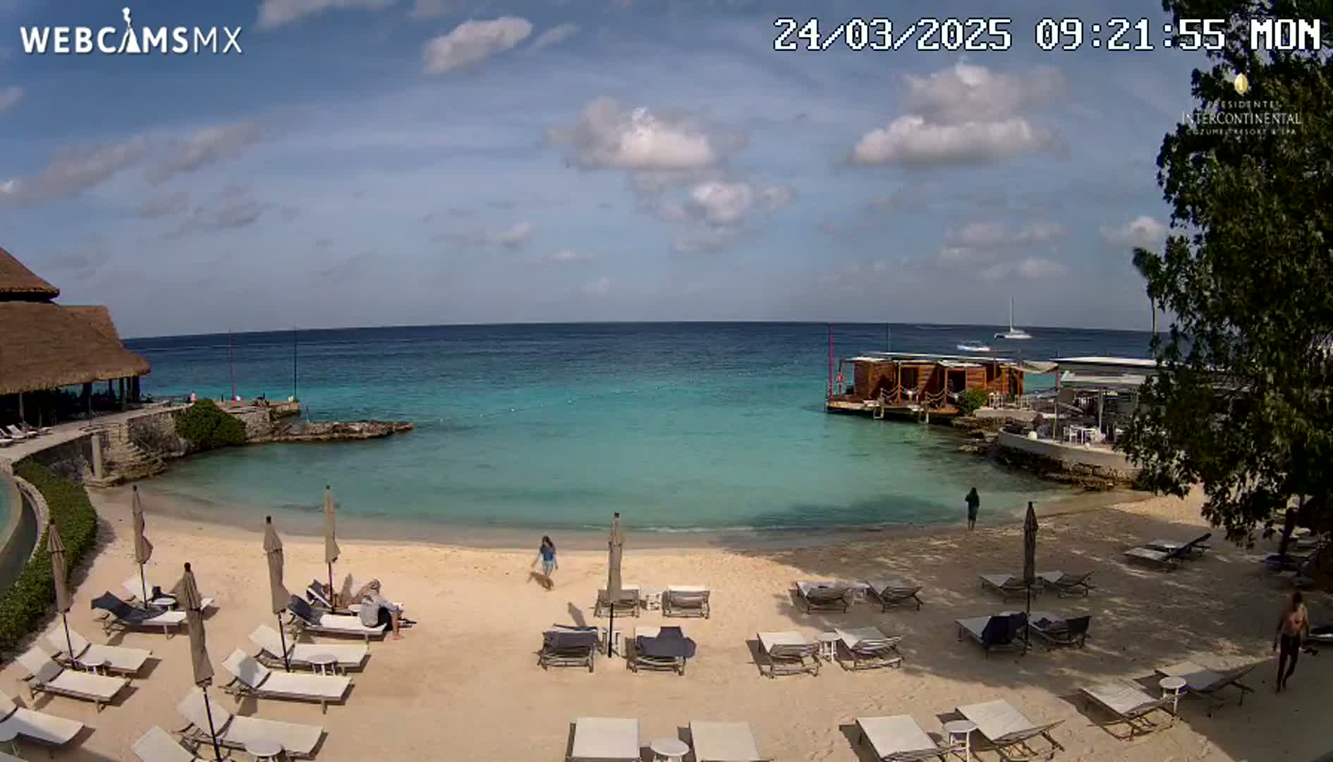 A sandy beach with lounge chairs and umbrellas sits beside a calm, turquoise ocean under a partly cloudy sky.