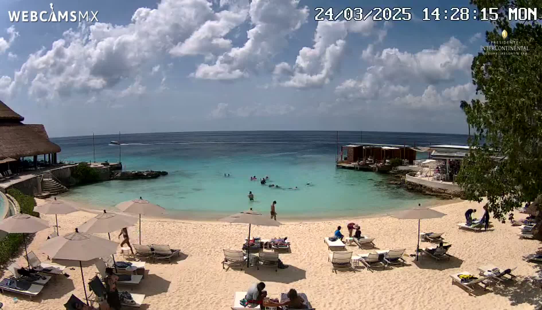 A partly sunny beach scene shows people relaxing on lounge chairs under umbrellas, others swimming in calm, clear turquoise water, and a thatched-roof building nearby.