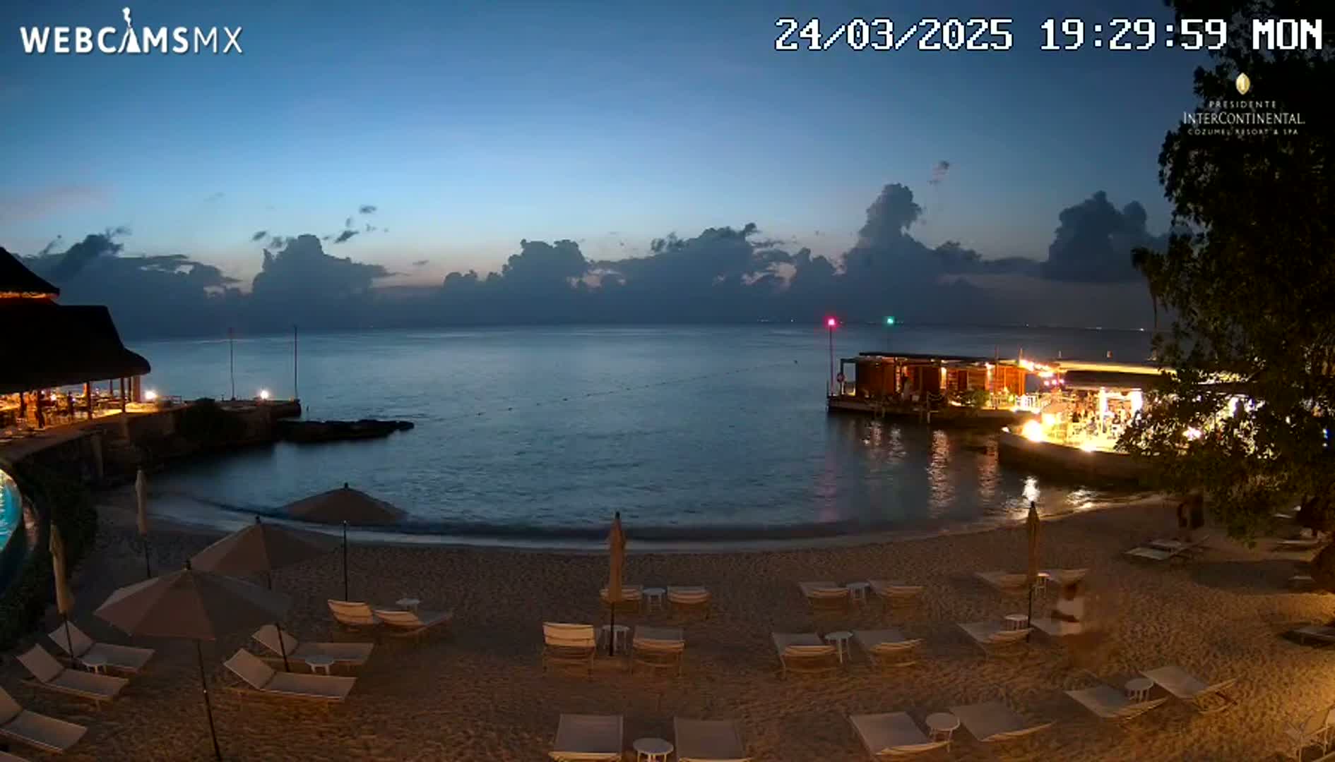 A beach at twilight with lounge chairs and umbrellas under a partly cloudy sky, showing a calm body of water with lights from an offshore structure.